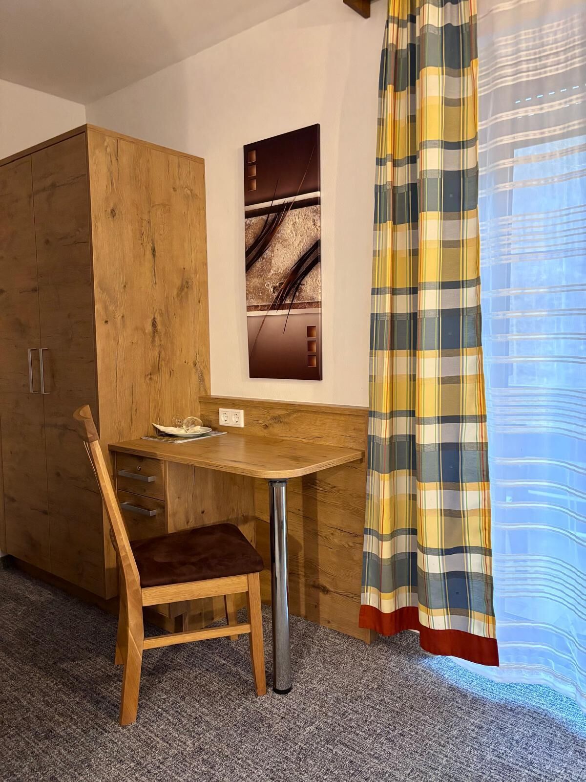 Um quarto pequeno com escrivaninha de madeira, uma cadeira, um guarda-roupa e cortina.