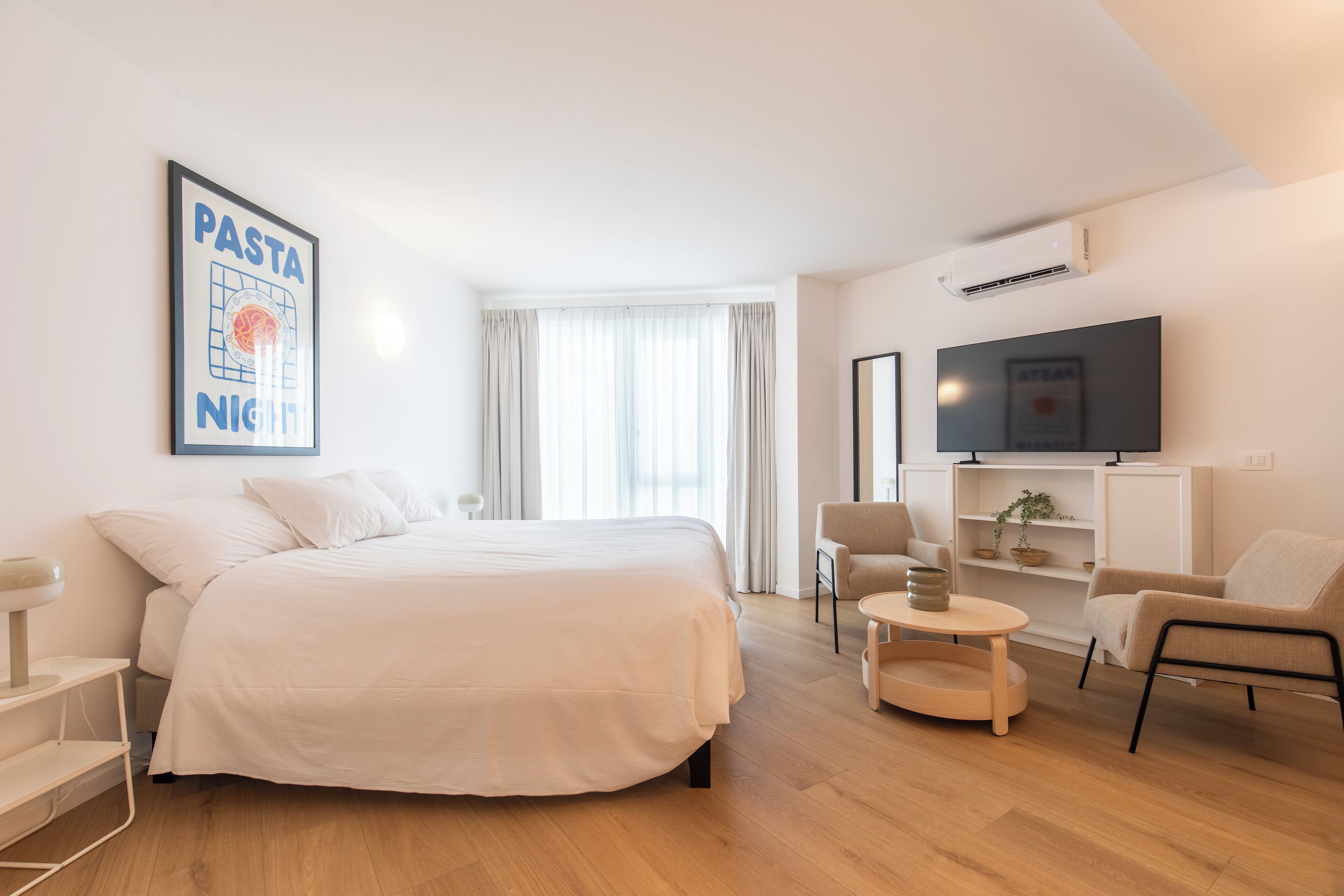 A modern hotel room with a large bed, a TV mounted on the wall, a small round table, and a chair.