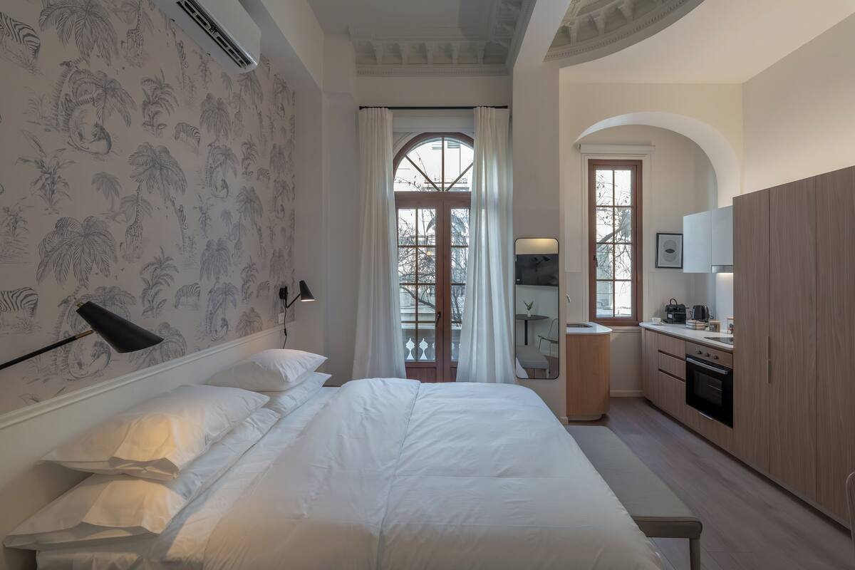 A bedroom with a large bed, a bedside table, a mirror, a window with curtains, and a kitchenette with cabinets and a stove.