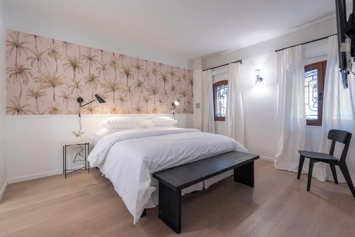 A bedroom with a bed, bedside table, lamp, bench, chair, and a wall with a palm tree pattern.