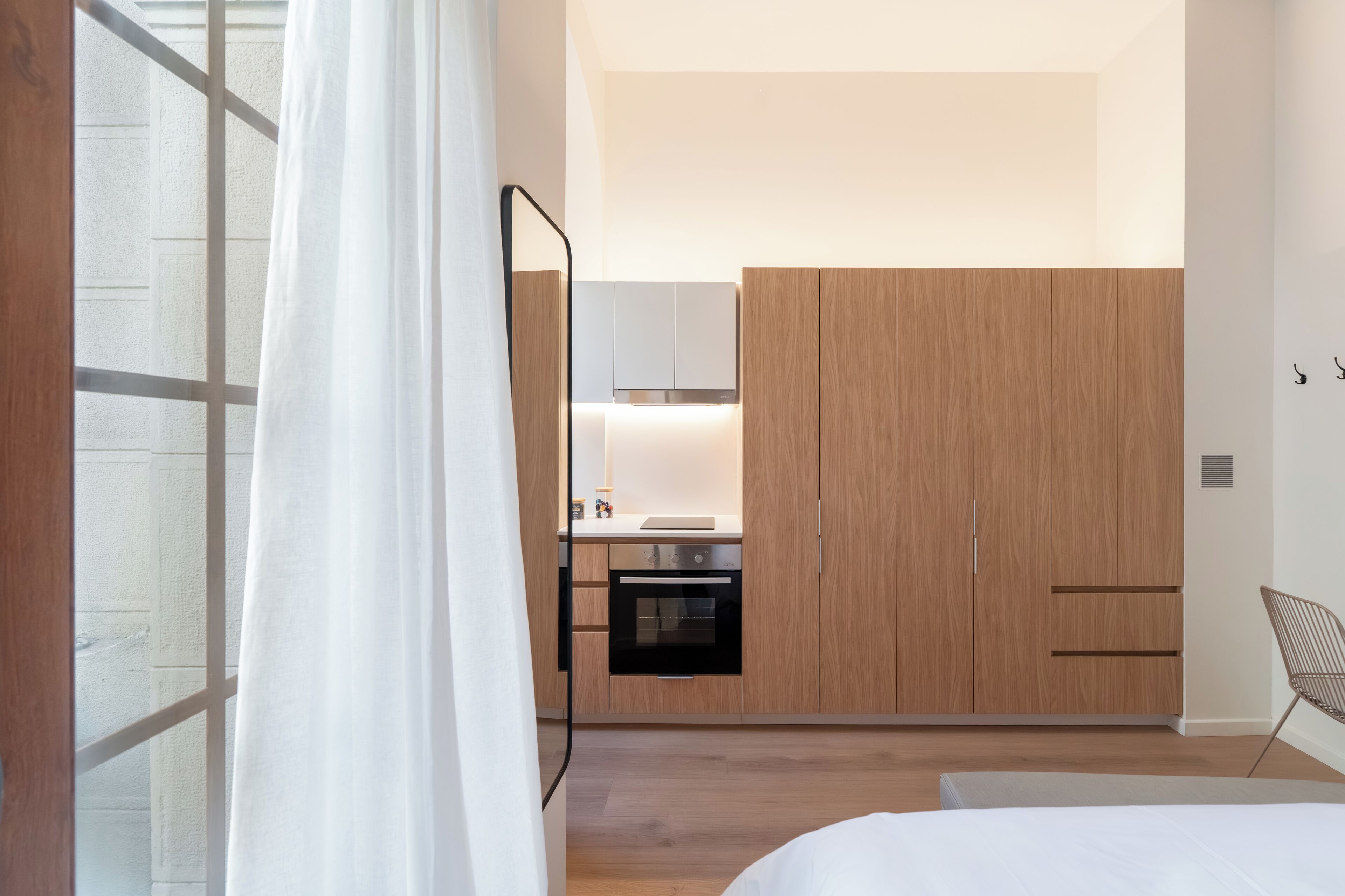 A modern bedroom with a large bed, a wooden wardrobe, a small kitchenette, and a window with curtains.