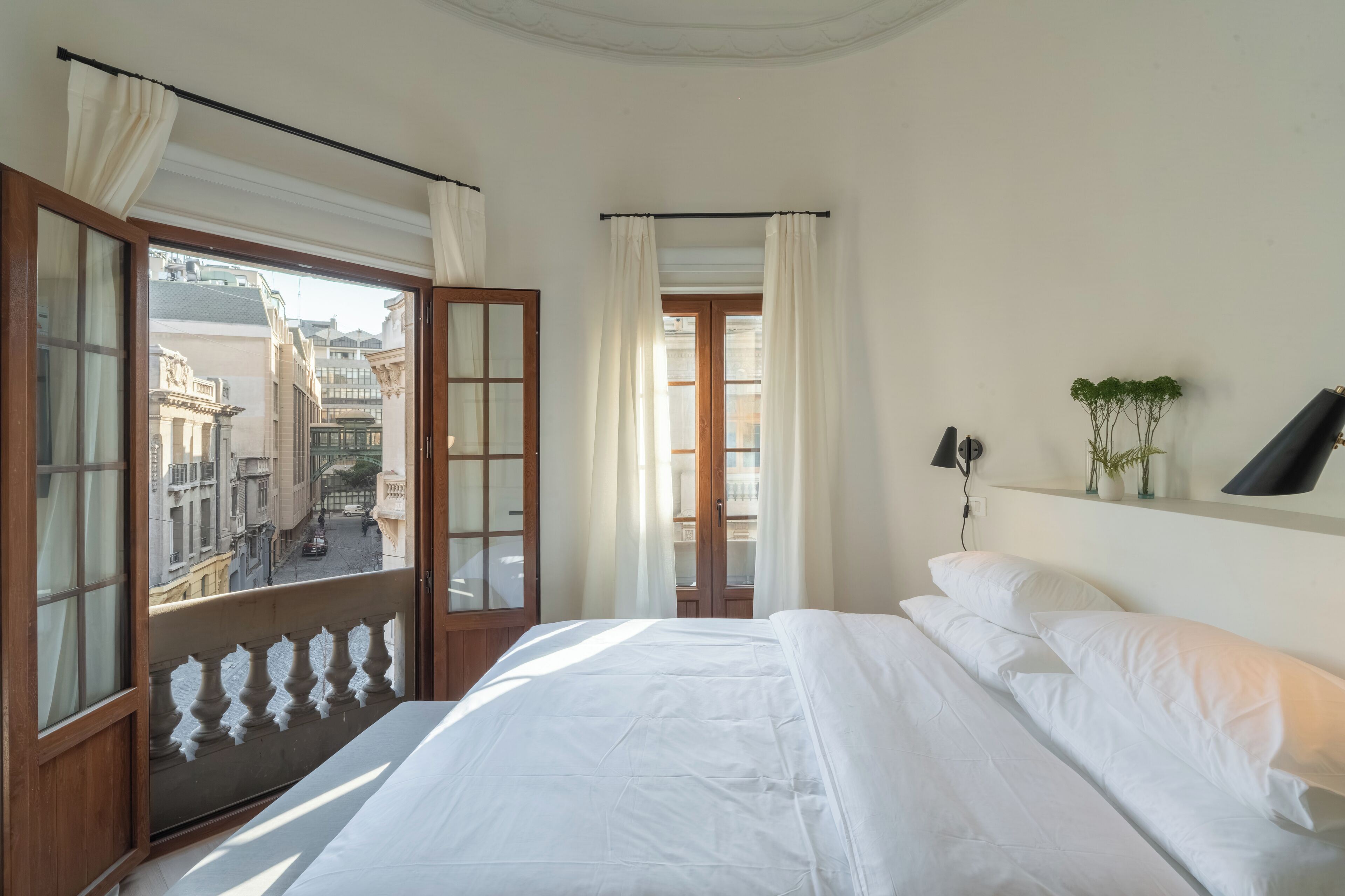 A bedroom with a large bed, a view of the city through a balcony door, and a small shelf with decorative items.
