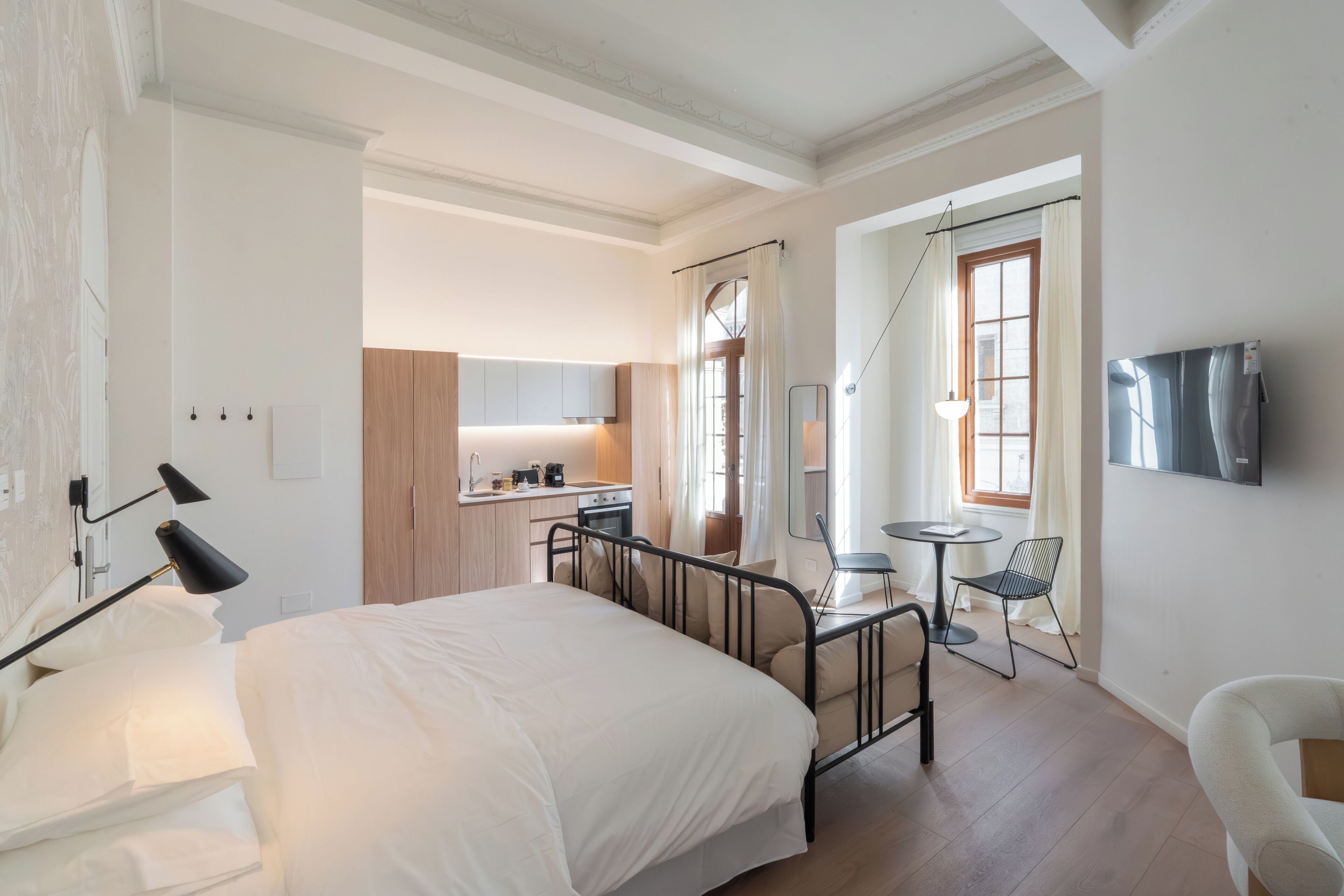 A modern bedroom with a large bed, a desk, and a chair, a small kitchenette, and a TV mounted on the wall.