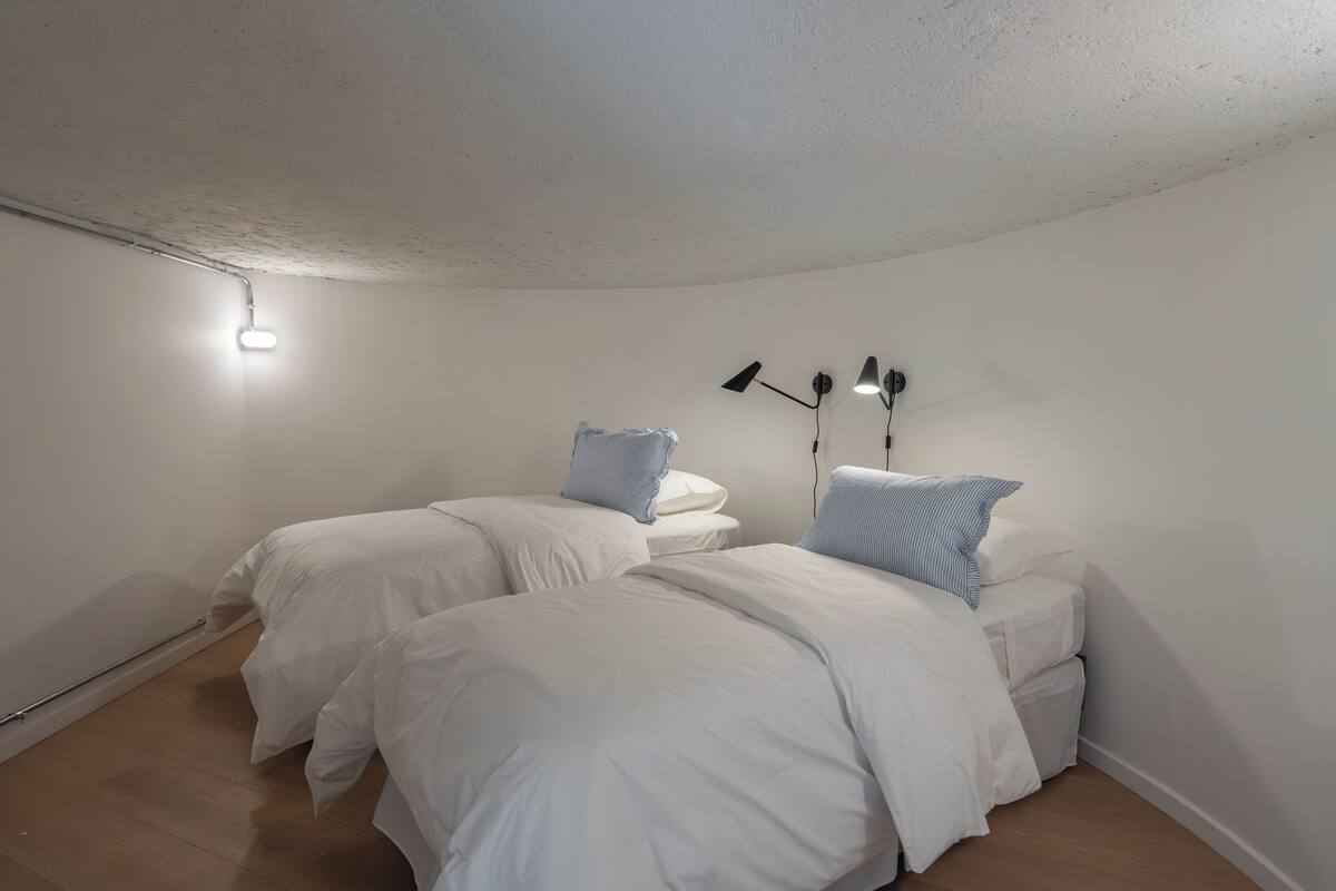 A bedroom with two beds, white bedding, a ceiling light, and a wall-mounted lamp.