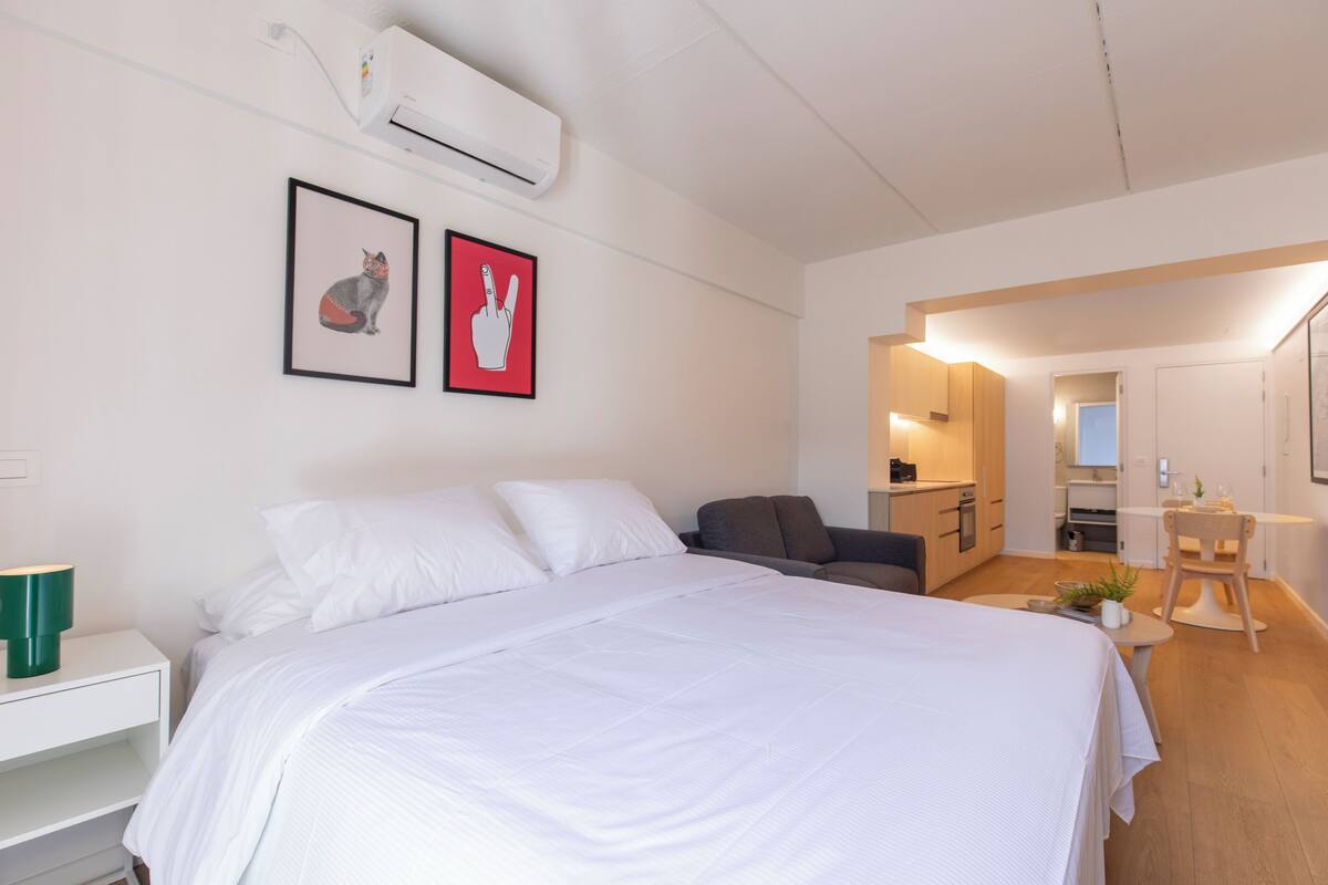 A modern hotel room with a large bed, a desk, and a kitchenette.