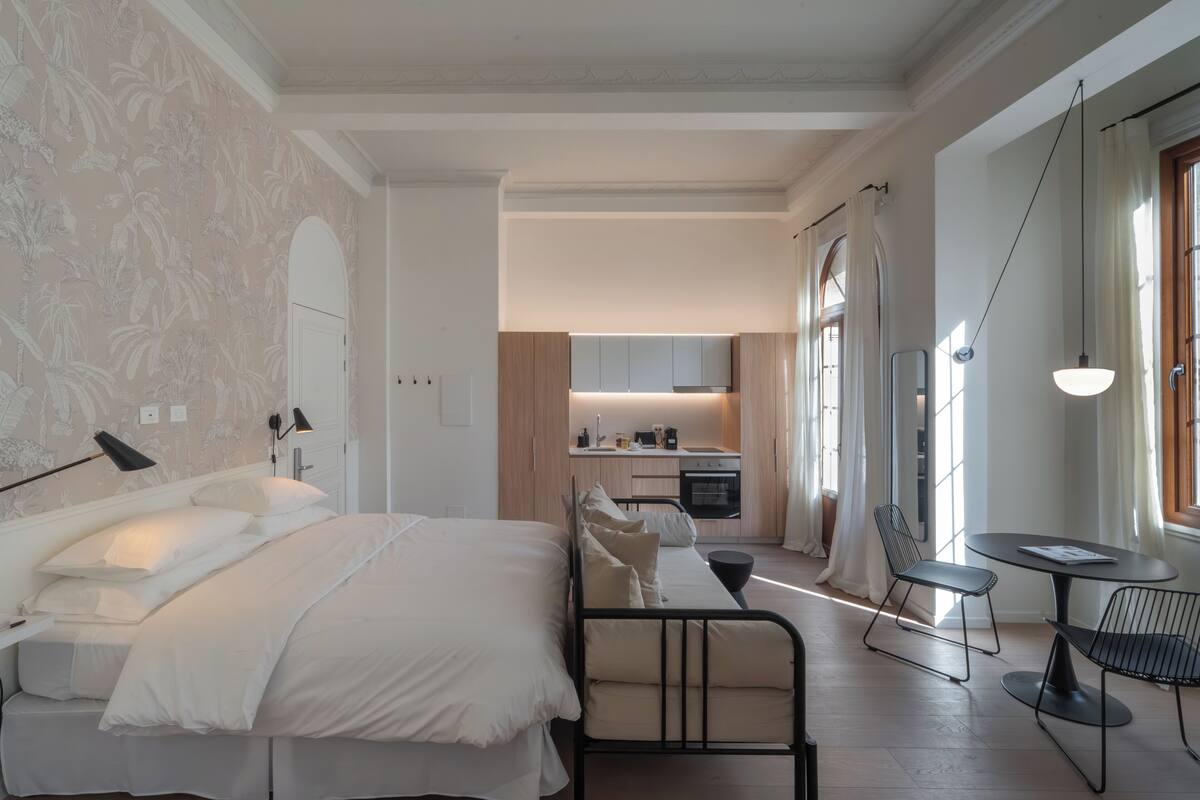 A modern hotel room with a large bed, a small kitchen area, and a dining table with chairs.