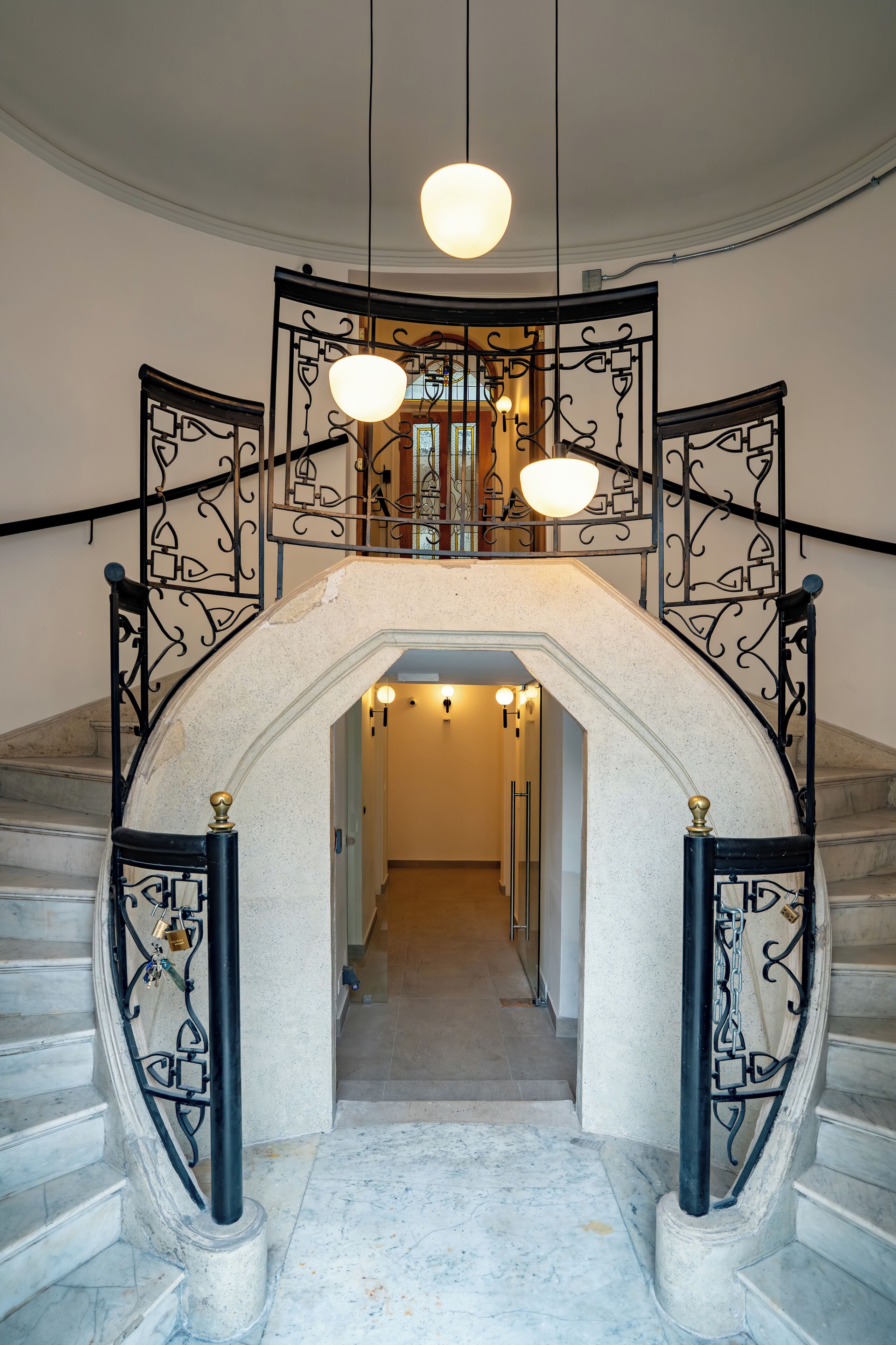 A grand staircase with ornate black railings and a marble floor, leading to a second-floor landing with a large, round light fixture.