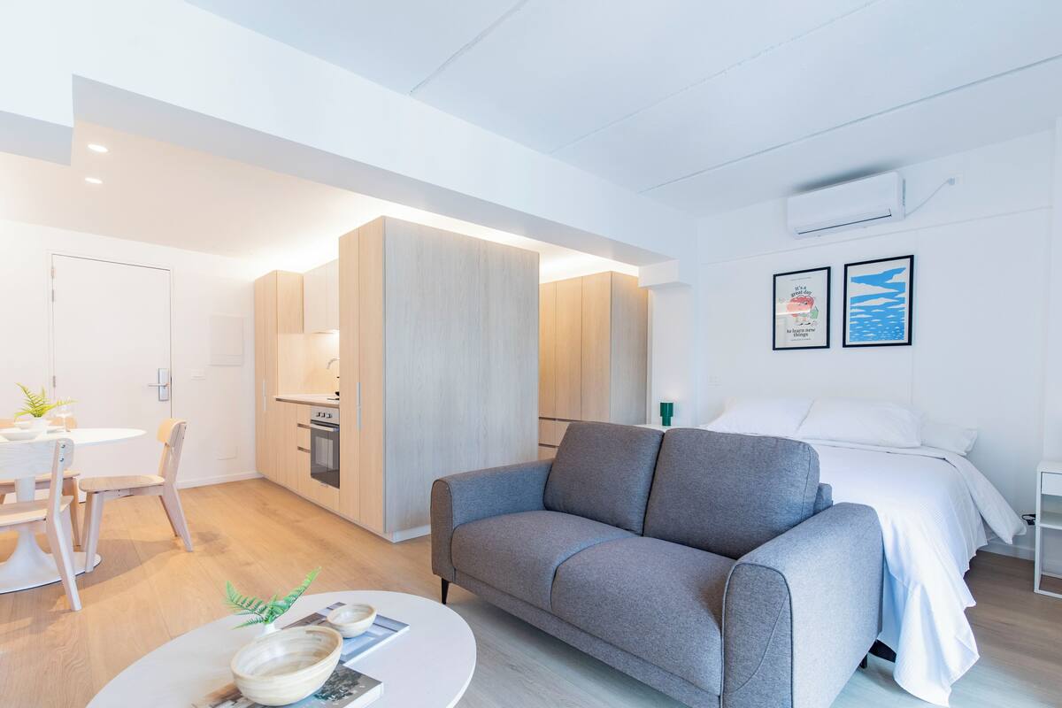 A modern, well-lit room with a grey sofa, a white bed, and a small kitchen area.