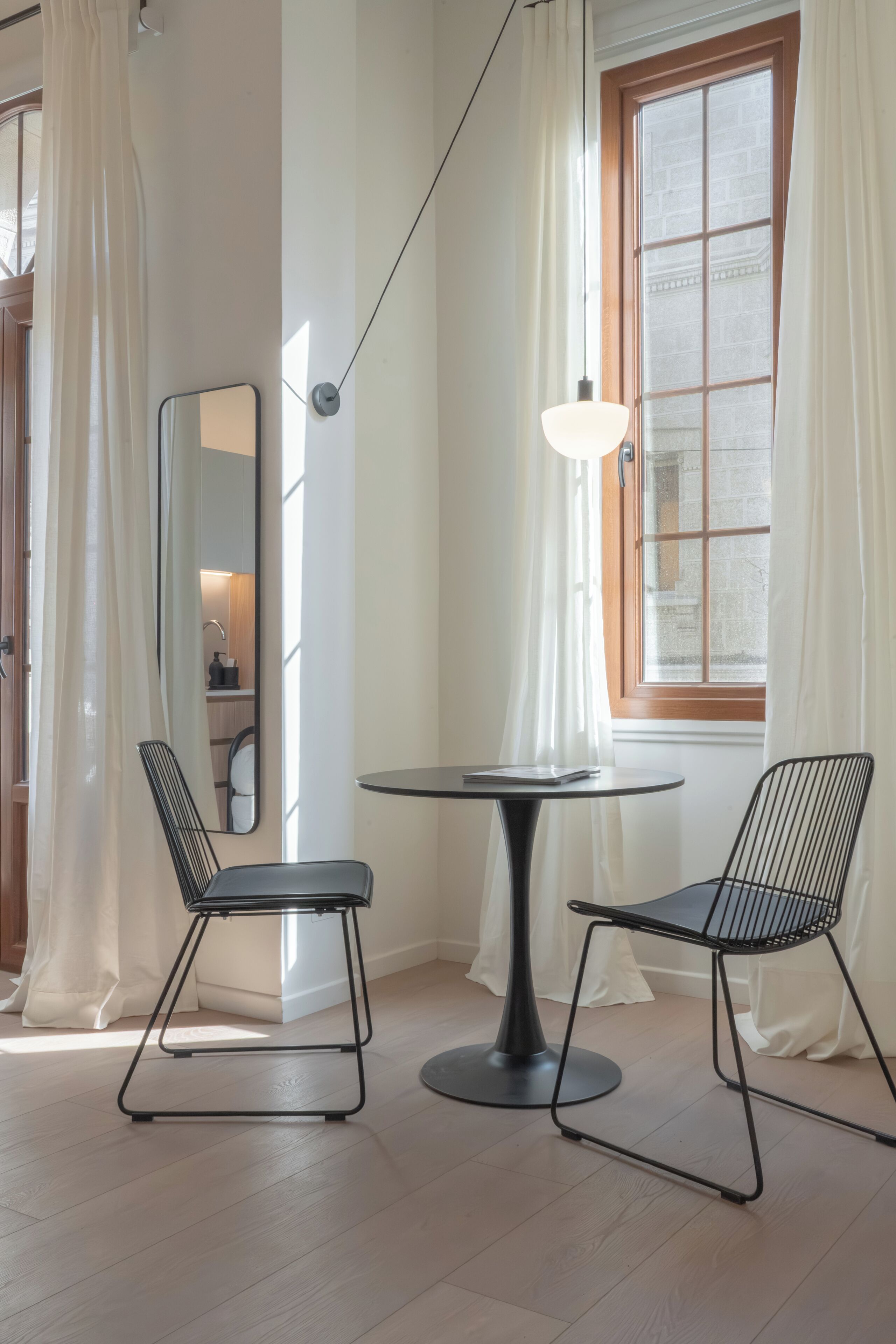A modern room with a round table, two black wireframe chairs, a floor lamp, a mirror, and a window with curtains.