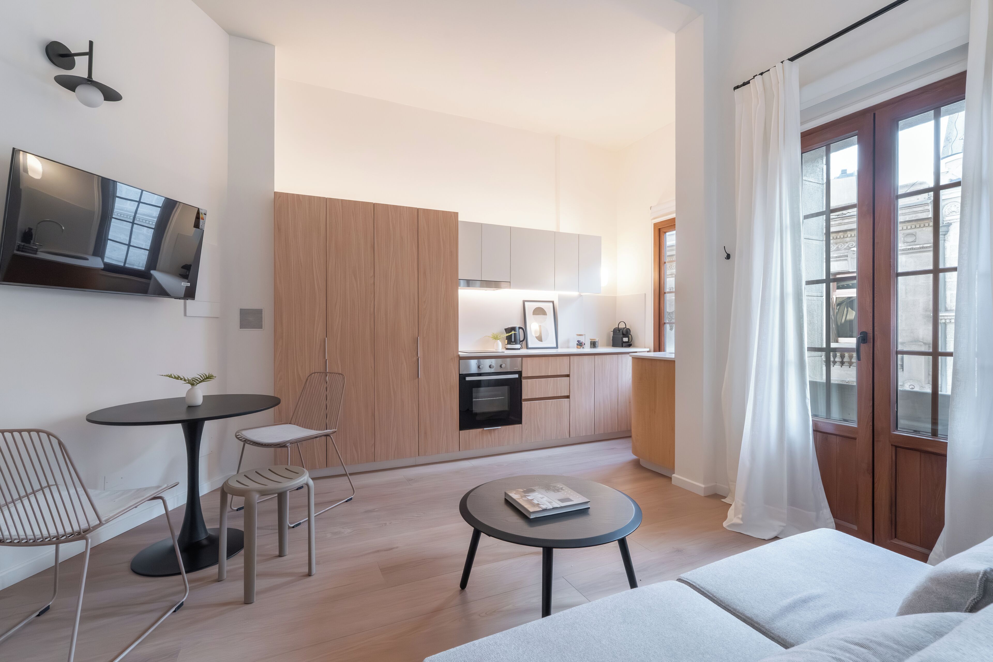 Ein modernes Wohnzimmer mit einer Couch, einem runden Couchtisch und einer kleinen Essecke mit Tisch und Stühlen. Im Hintergrund ist eine Küche mit Holzschränken und einem eingebauten Ofen zu sehen.
