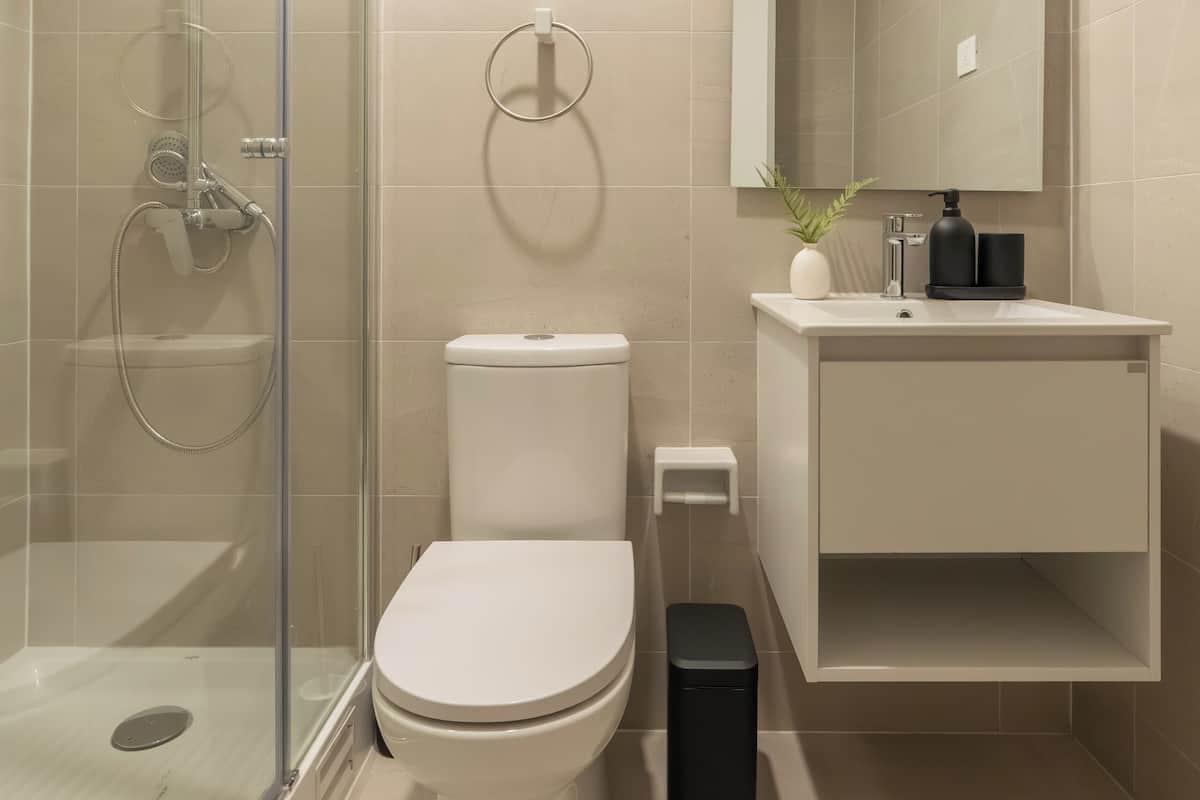 A modern bathroom with a glass-enclosed shower, a toilet, a sink with a mirror, and a trash bin.