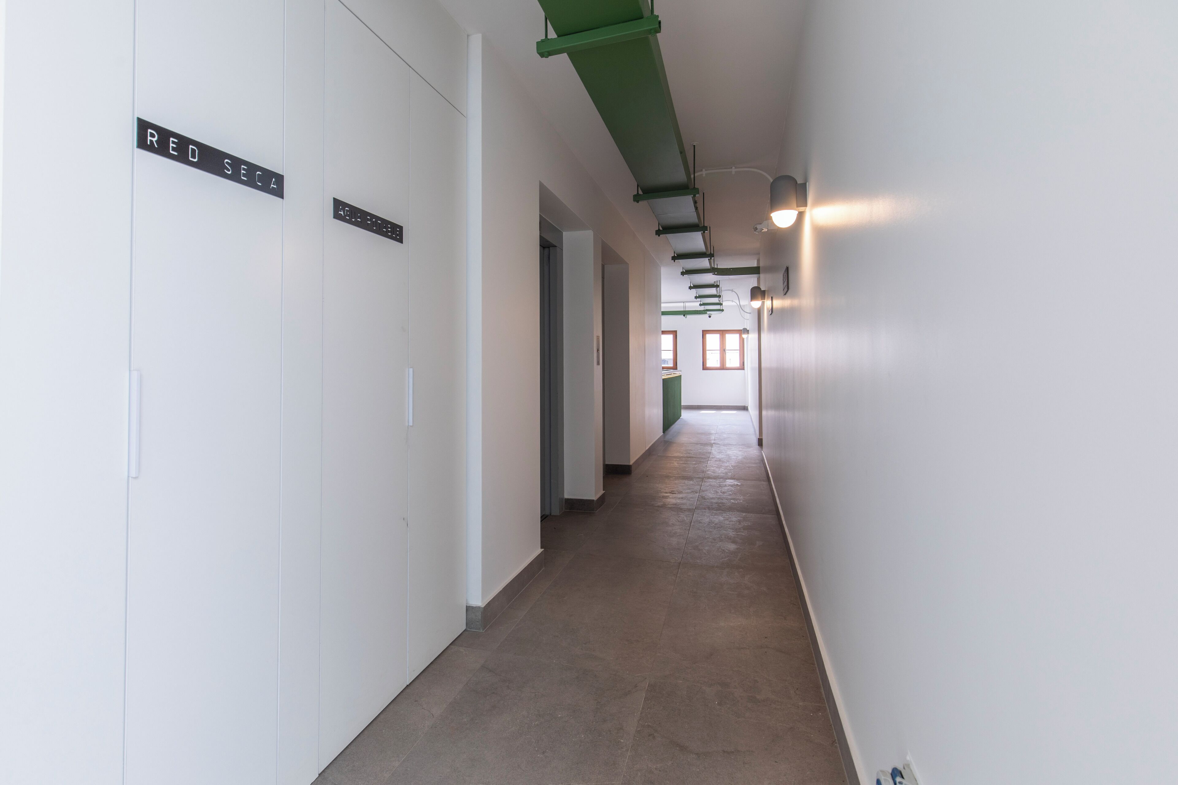 Un corridoio con pareti bianche, pavimento marrone e tubi al soffitto verdi. Ci sono cartelli appesi alle pareti e un lampadario sulla destra.