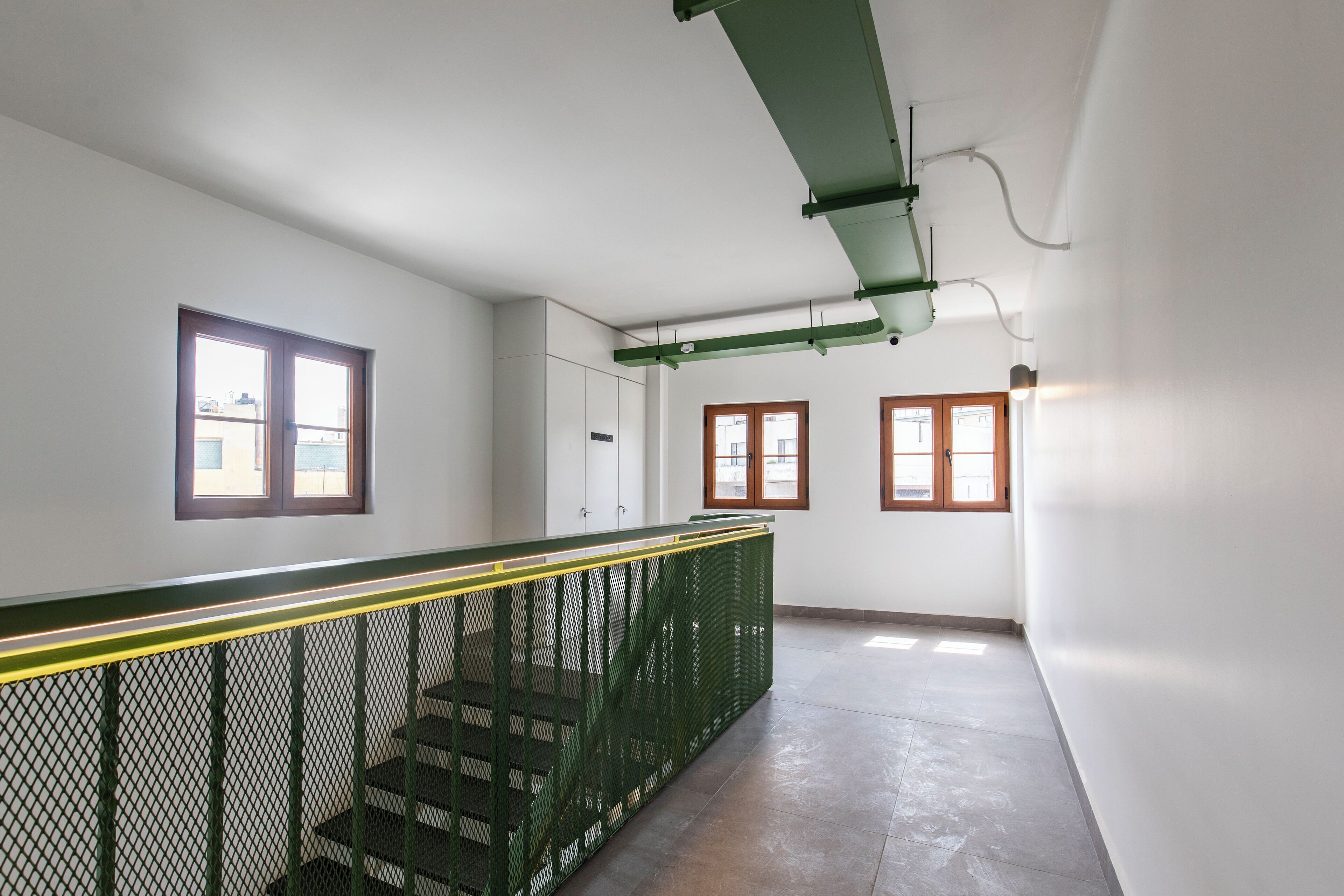 A staircase with green railings and a yellow safety line, leading to a landing with a white wall and a window with a wooden frame.