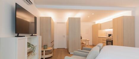 A modern hotel room with a flat-screen TV, a sofa, a bed, a small table, and a kitchenette.