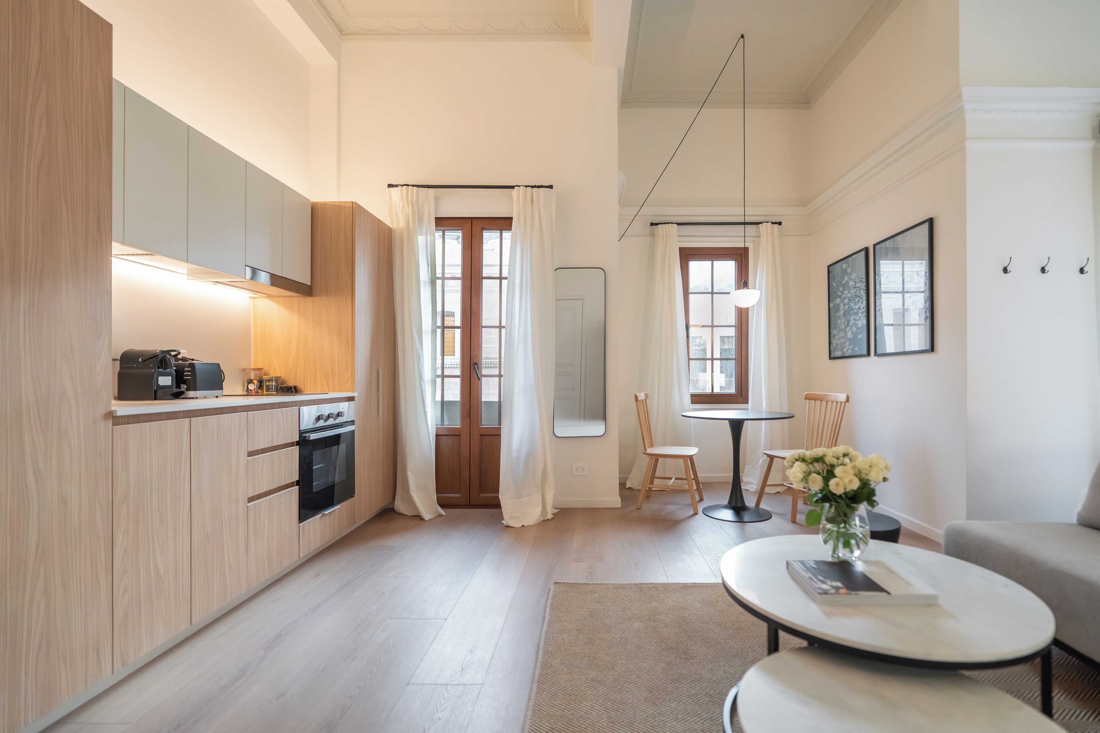 Una cocina moderna con armarios de madera, horno integrado y máquina de café. Hay un comedor con mesa y sillas, y una sala de estar con sofá y mesa de centro.