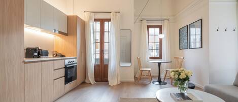 Una cocina moderna con armarios de madera, horno integrado y máquina de café. Hay un comedor con mesa y sillas, y una sala de estar con sofá y mesa de centro.
