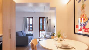 Una sala de estar moderna con comedor, un sofá, una mesa con un florero y copas de vino, y un ventanal con cortinas.