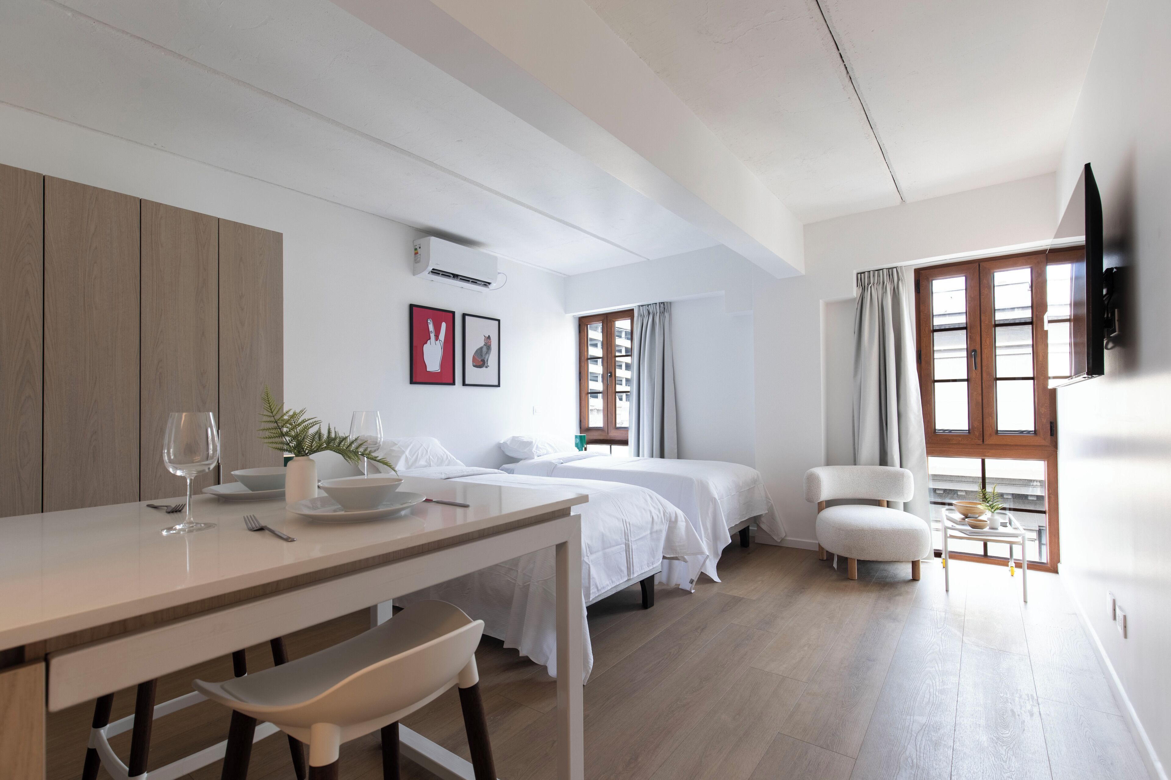 Una habitación moderna y bien iluminada con una mesa de comedor de madera, dos camas blancas y una pequeña zona de estar.