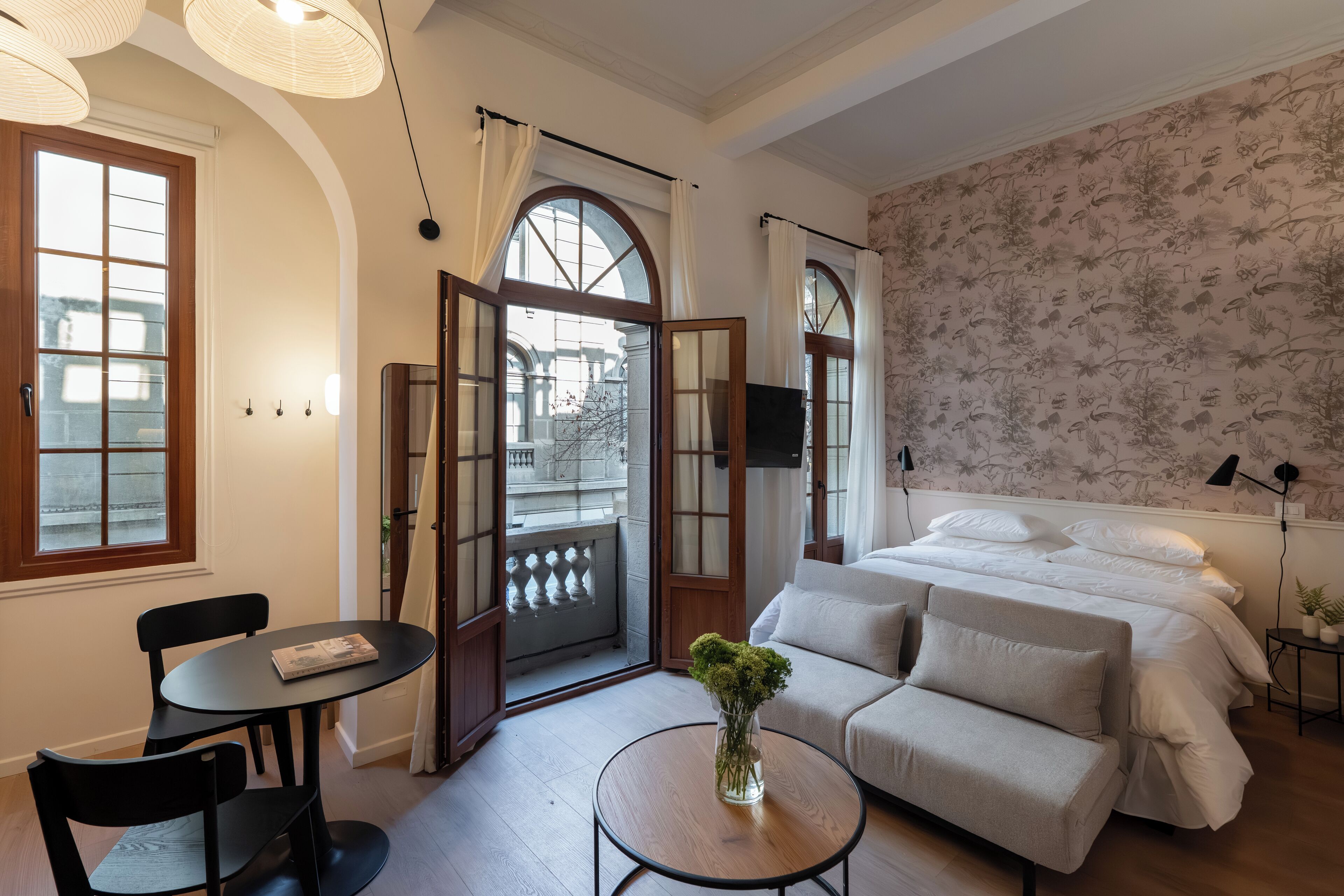 A modern bedroom with a bed, a sofa, a small table with a vase, and a dining area with chairs.