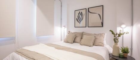 A modern bedroom with a large bed, a bedside table with a lamp, and two framed artworks on the wall.