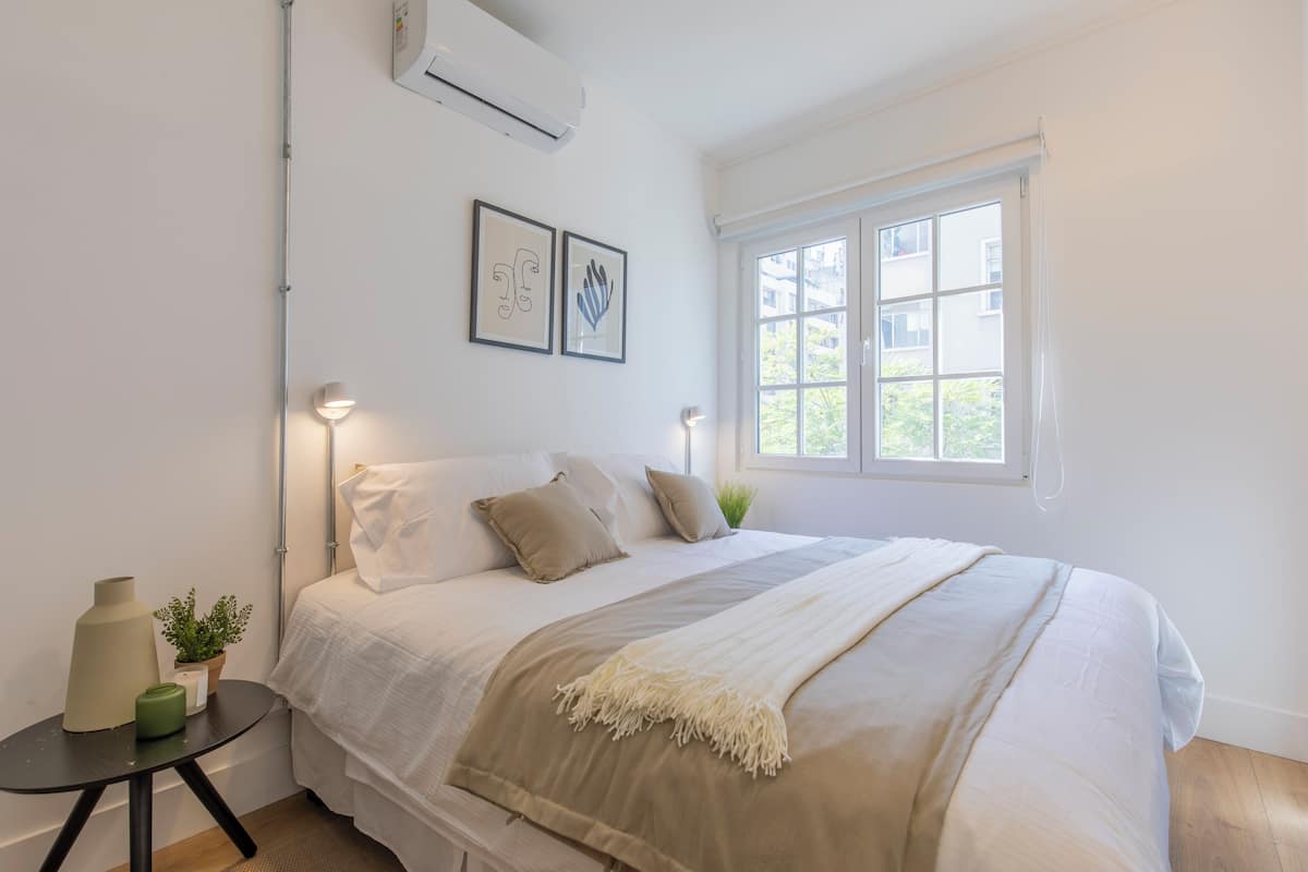 Un dormitorio con cama, mesitas de noche, ventana y aire acondicionado instalado en la pared.