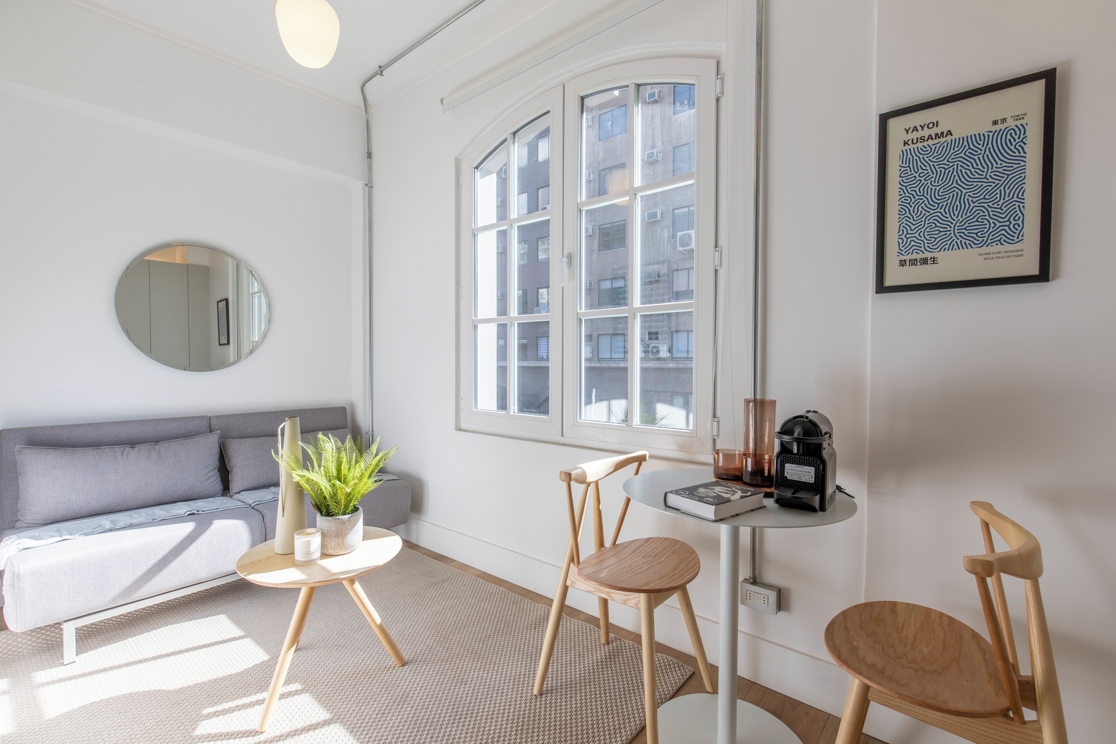 A modern living room with a grey sofa, a round mirror, a small round table with a plant, two wooden chairs, and a coffee machine on a small table.