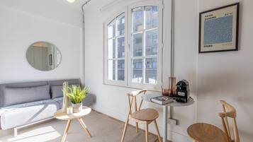 A modern living room with a grey sofa, a round mirror, a small round table with a plant, two wooden chairs, and a coffee machine on a small table.