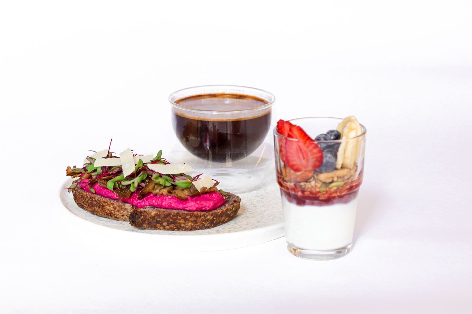 A plate with a beetroot and cheese sandwich, a cup of coffee, and a glass of yogurt with berries and granola.