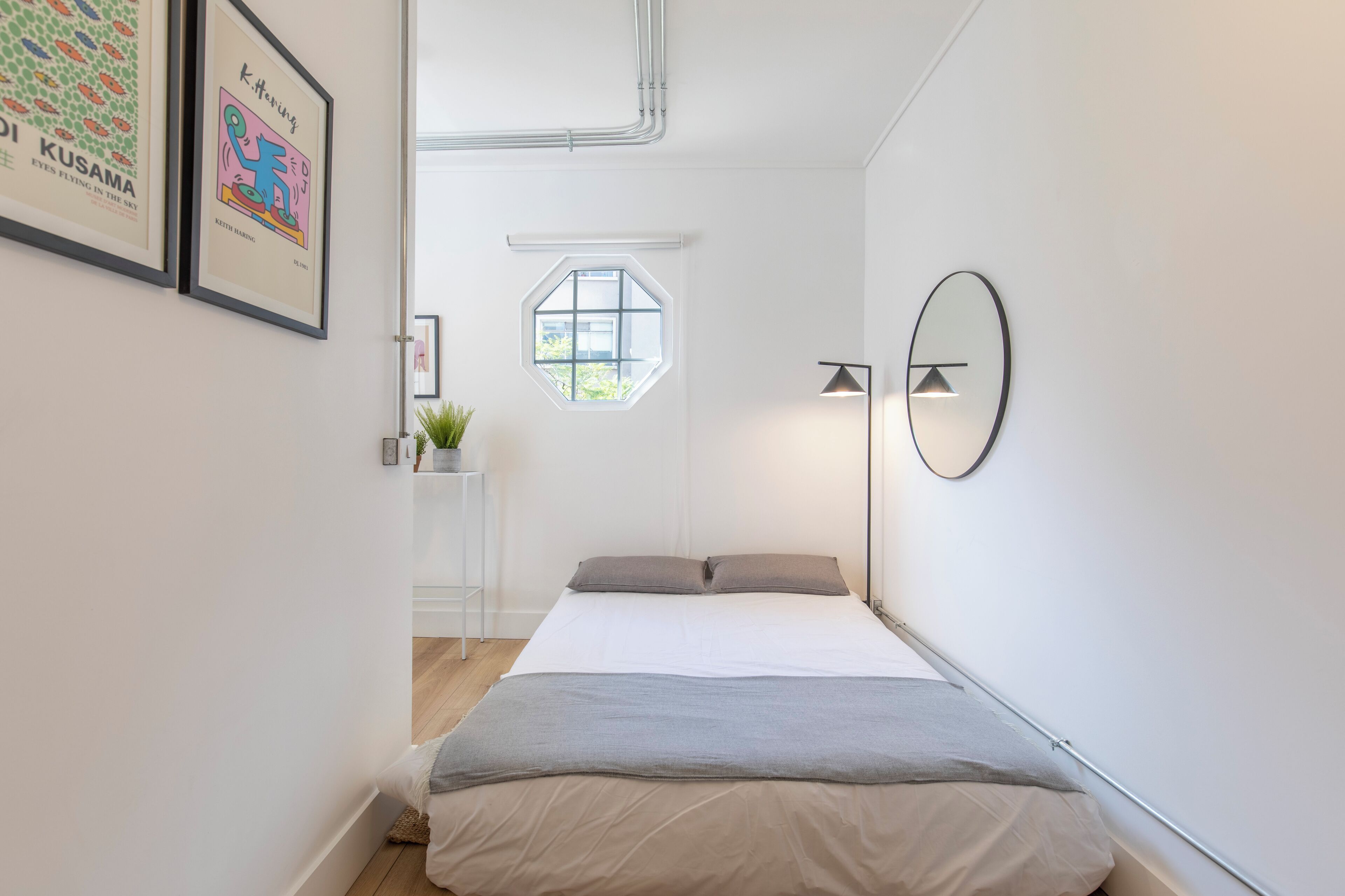 A bedroom with a bed, a window, a wall-mounted mirror, and framed artwork.