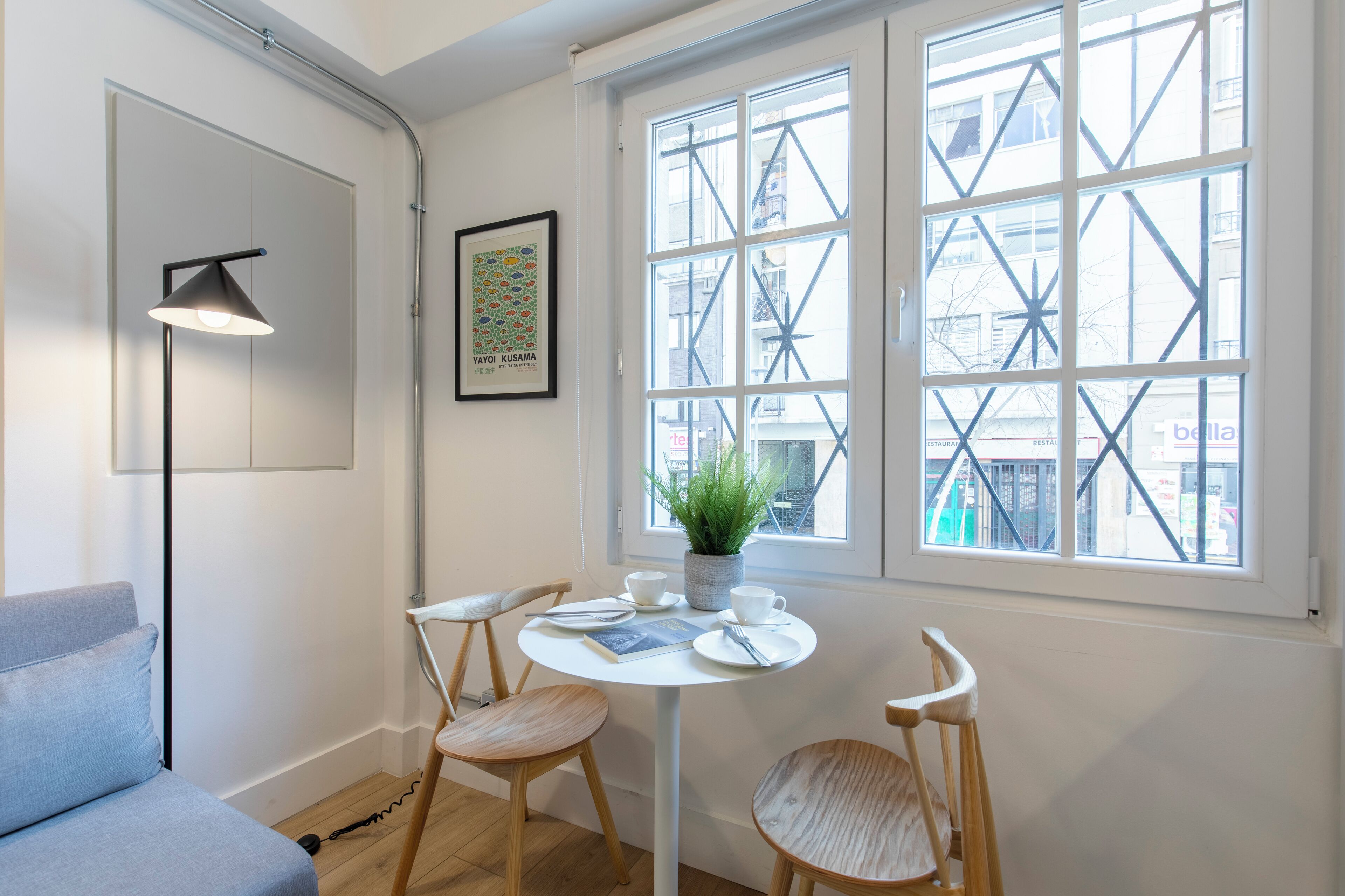 A room with a white interior, a round table with chairs, a floor lamp, a potted plant, and a window with a view of buildings.