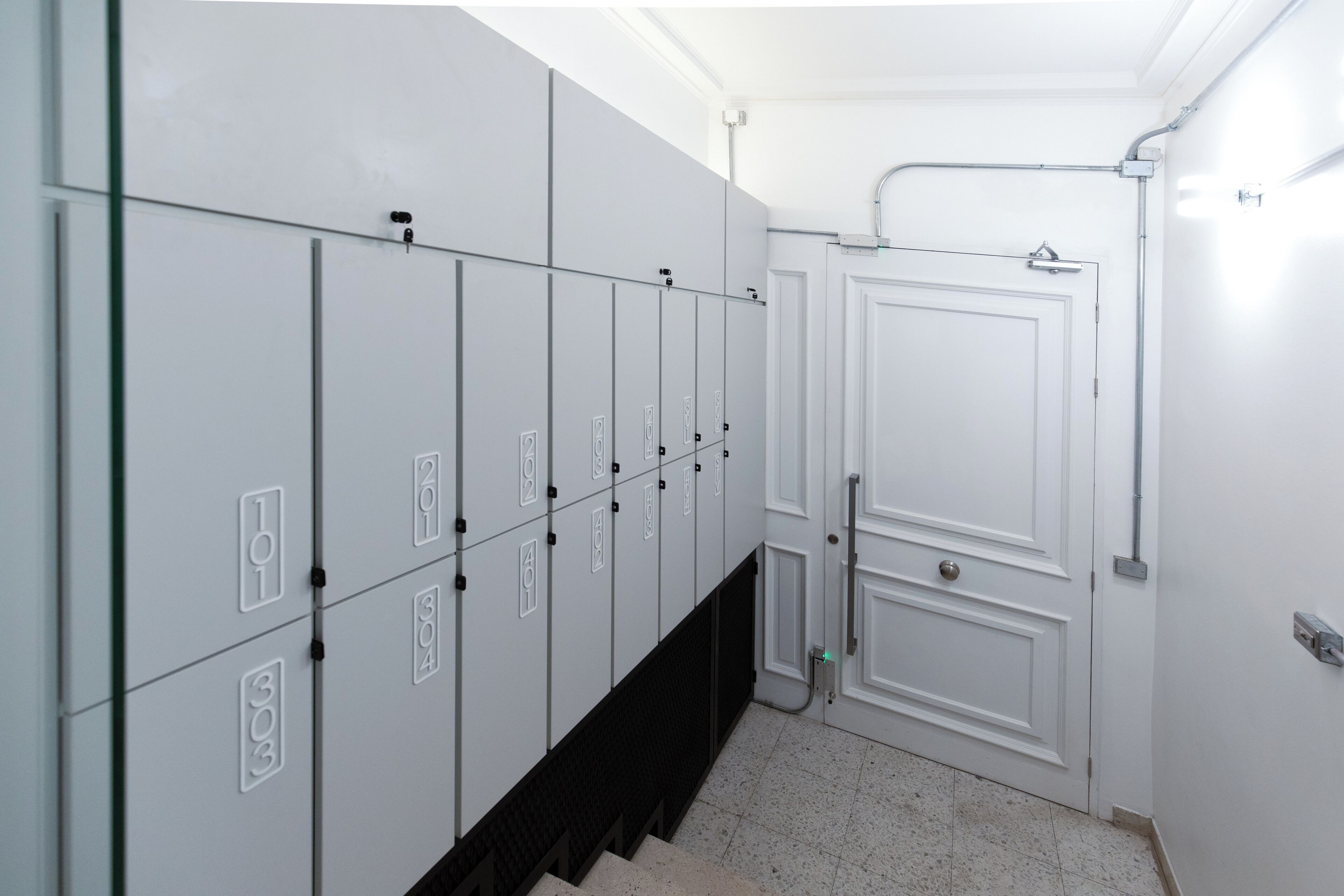 Un pasillo con casilleros numerados del 101 al 303, una puerta blanca y piso de baldosas.
