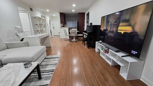 A modern living room with a large flat-screen TV, a piano, a dining area, and a kitchen in the background. - Woodland Hill Condo with Pool, Gym, Free Parking (Los Angeles)