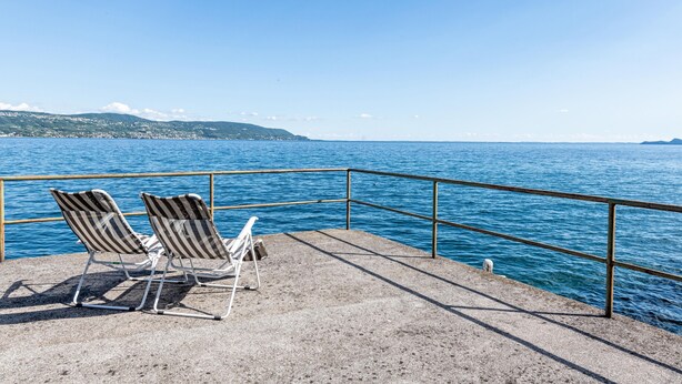 Una terraza de hormigón con dos sillones de exterior a rayas, orientados hacia una masa de agua, una barandilla metálica y una vista distante de la costa.