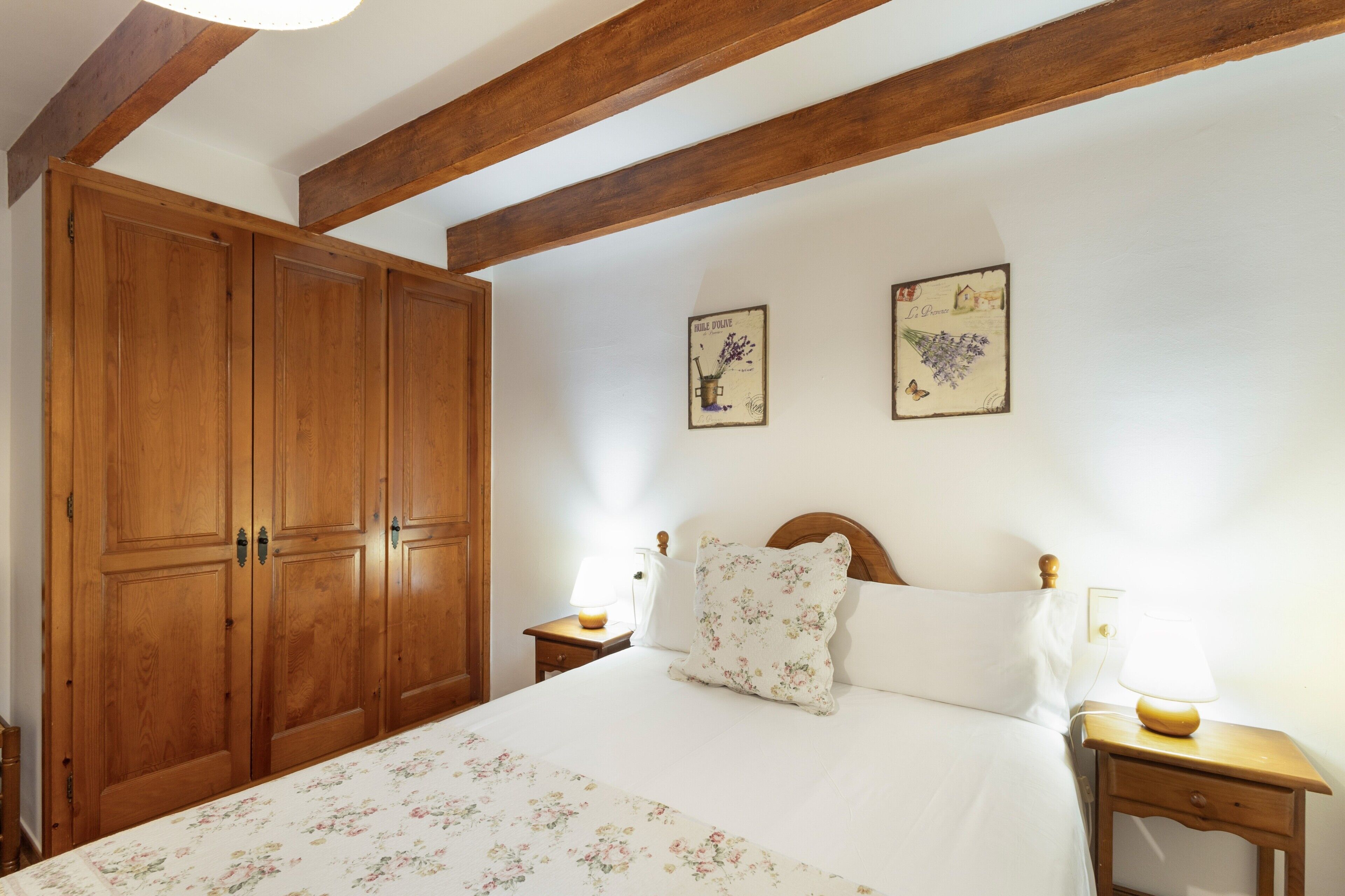 Een slaapkamer met een grote houten garderobe, een bed met bloemenbeddengoed, twee nachtkastjes met lampen en twee ingelijste foto’s aan de muur.