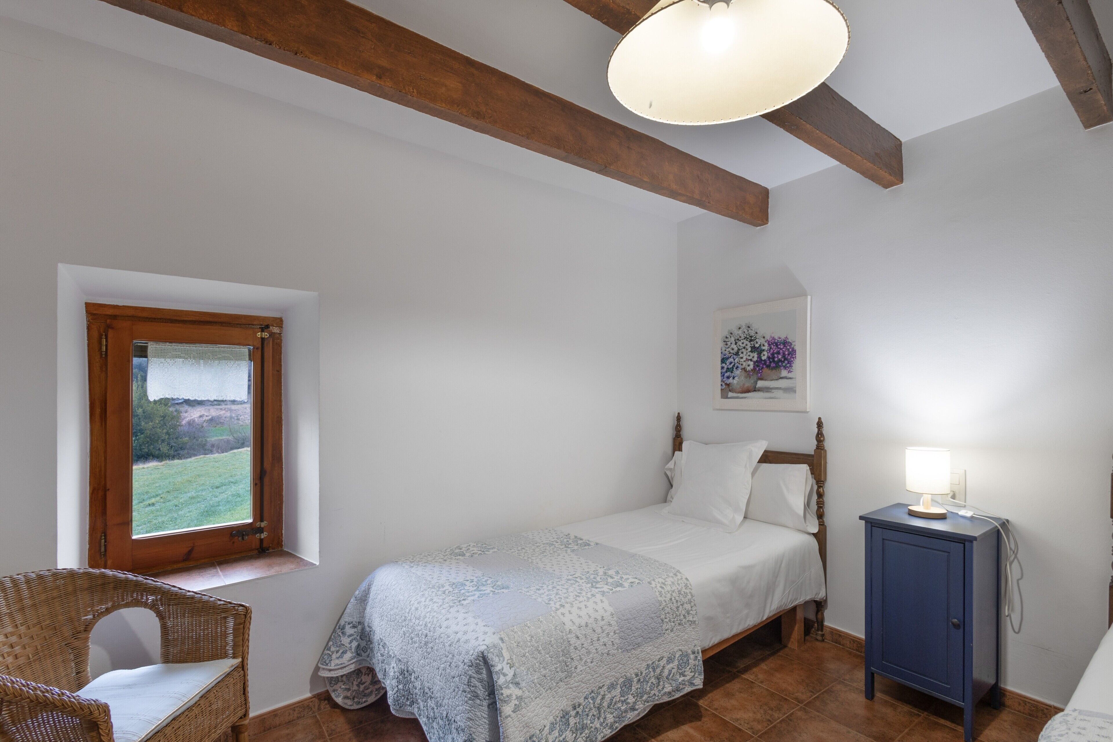 A bedroom with a bed, a nightstand with a lamp, a chair, a window with a view, and a wooden ceiling beam.