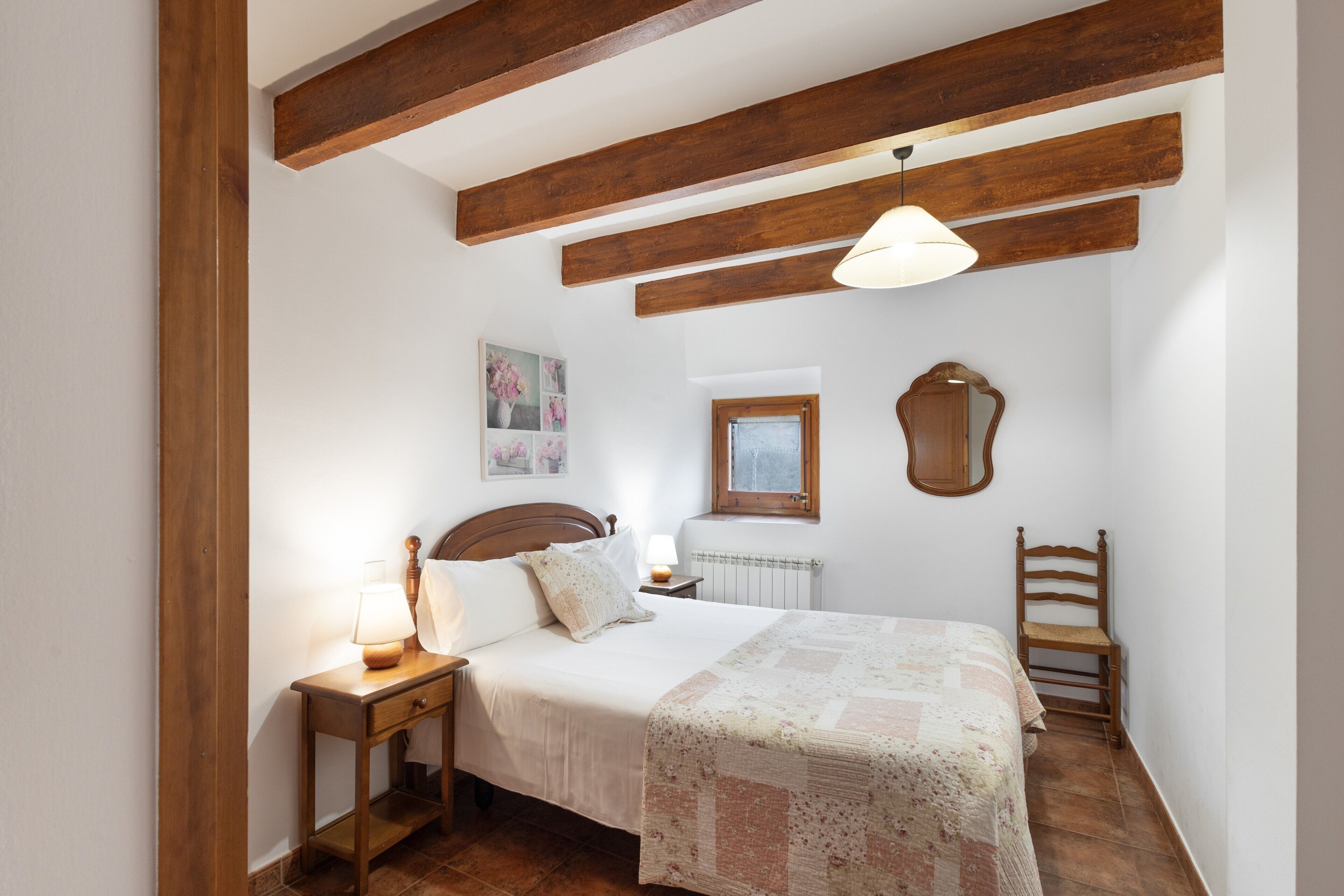 A bedroom with a wooden ceiling, a bed with a quilt, a nightstand with a lamp, a chair, and a wall-mounted mirror.