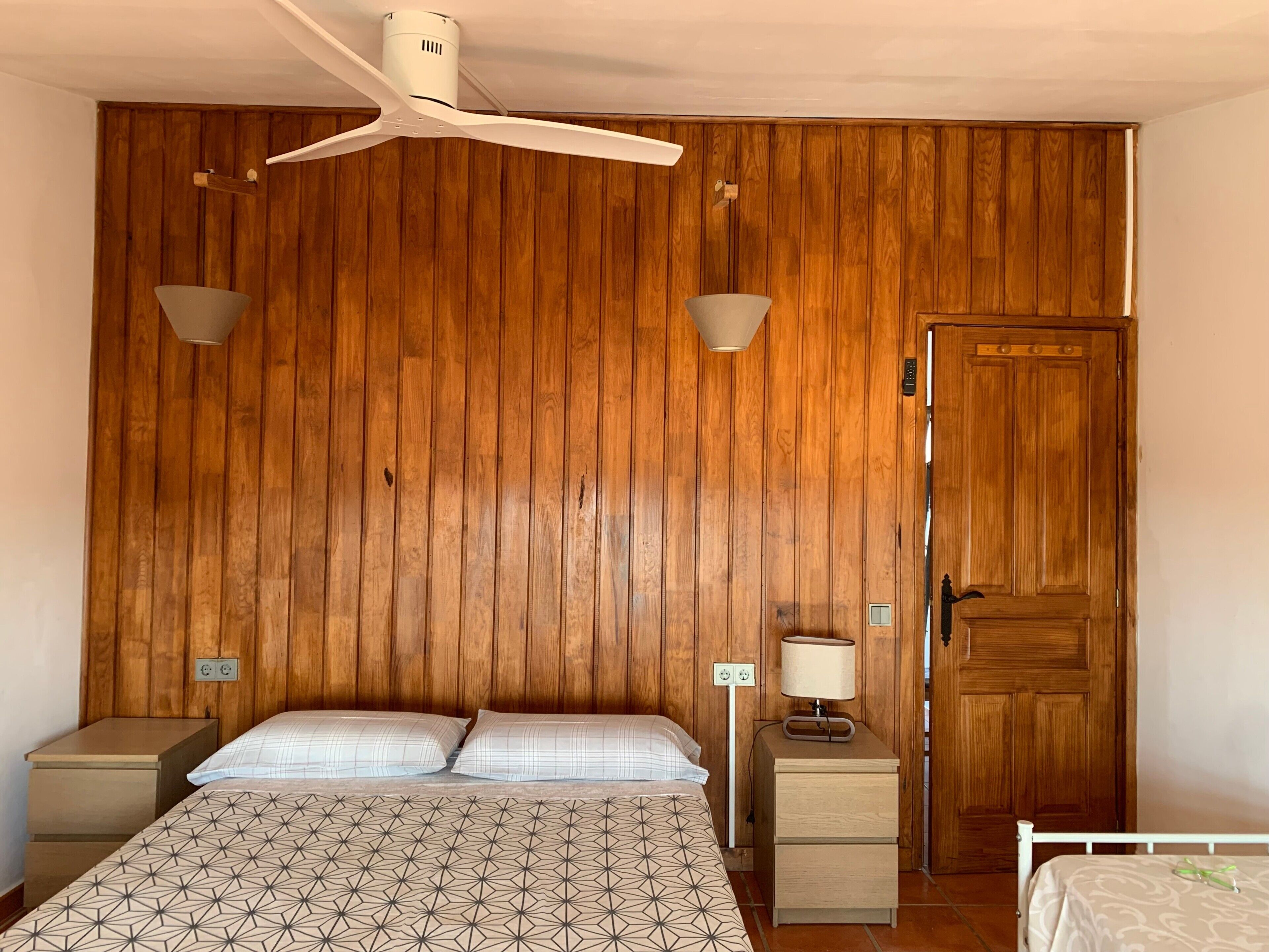 Un dormitorio con pared de paneles de madera, una cama con ropa de cama estampada, un ventilador de techo, lámparas de noche, una mesita de noche y una puerta.