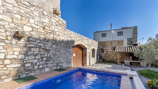 A stone building with a swimming pool, a wooden door, and a small garden area.