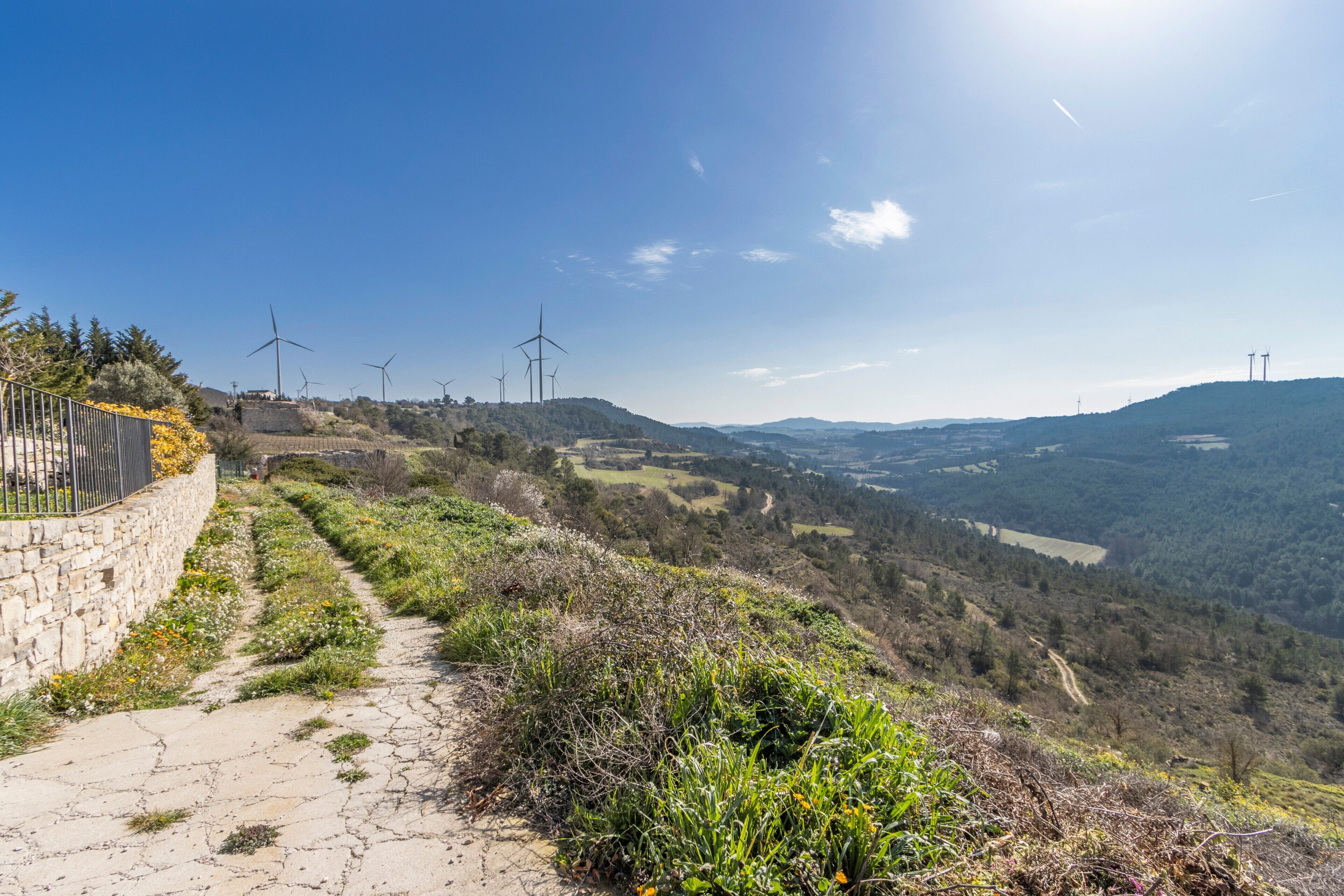 Un sentiero che conduce verso un paesaggio collinare con turbine eoliche e un muro in pietra a sinistra.