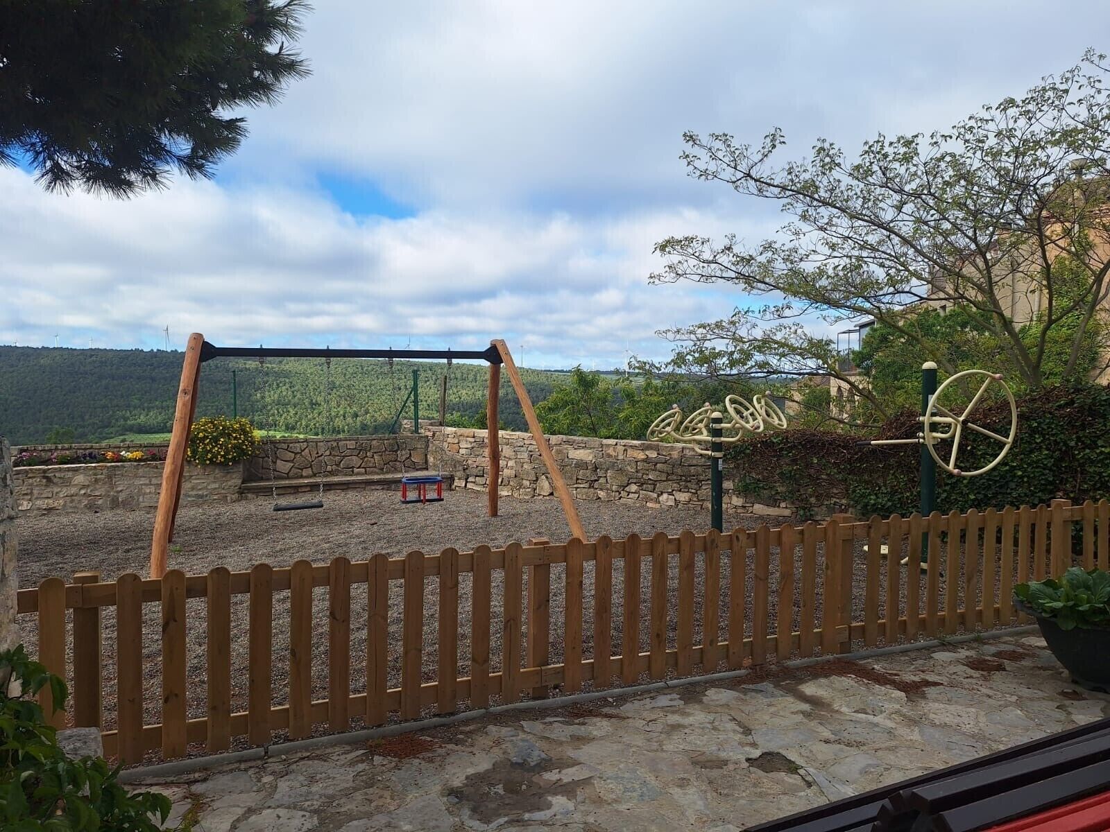A playground with swings and a merry-go-round, surrounded by a wooden fence and a stone wall.