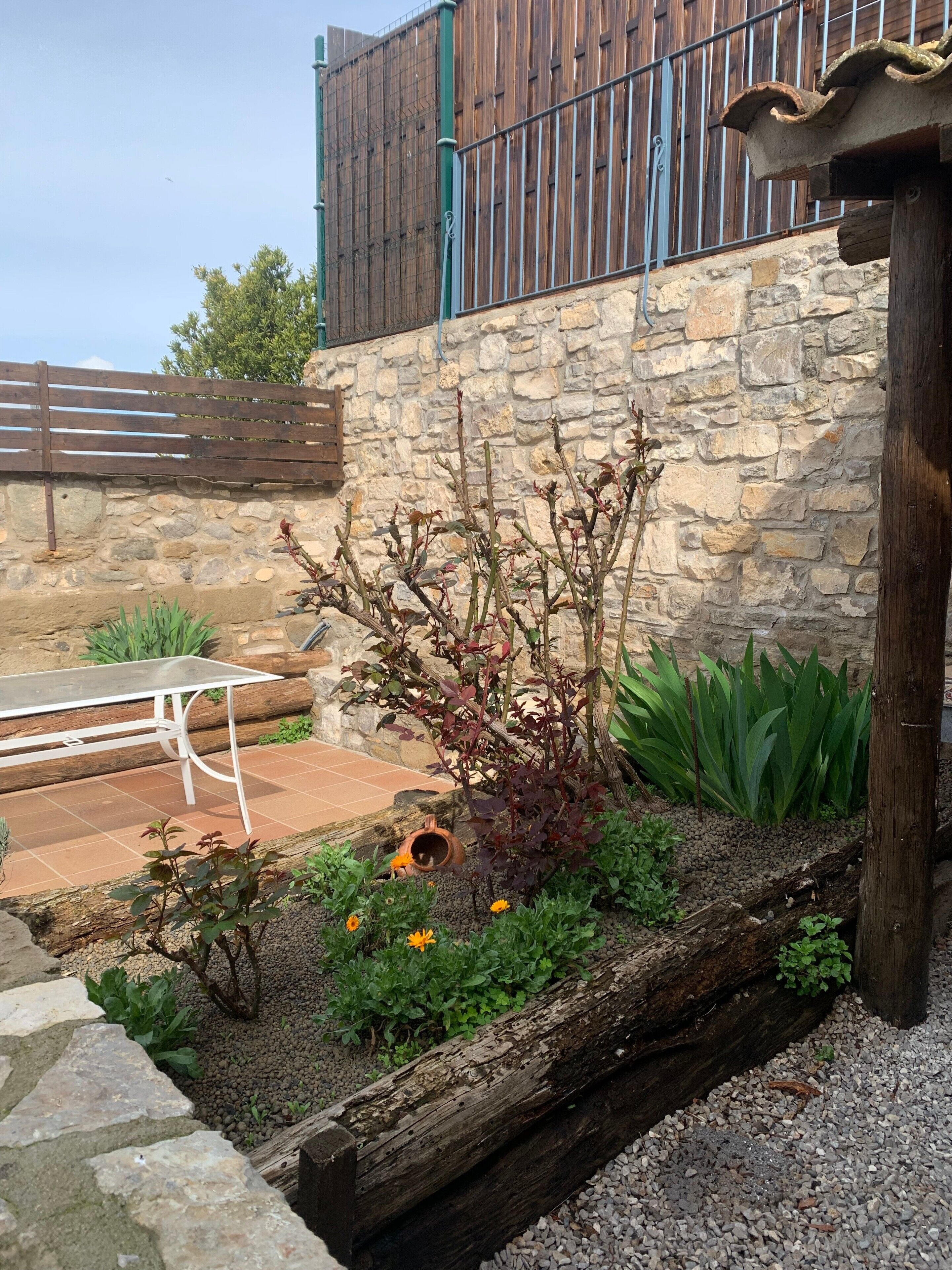 Un muro de piedra, una cerca de madera, una mesa de metal y un jardín con plantas y flores.