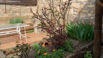 Un muro de piedra, una cerca de madera, una mesa de metal y un jardín con plantas y flores.