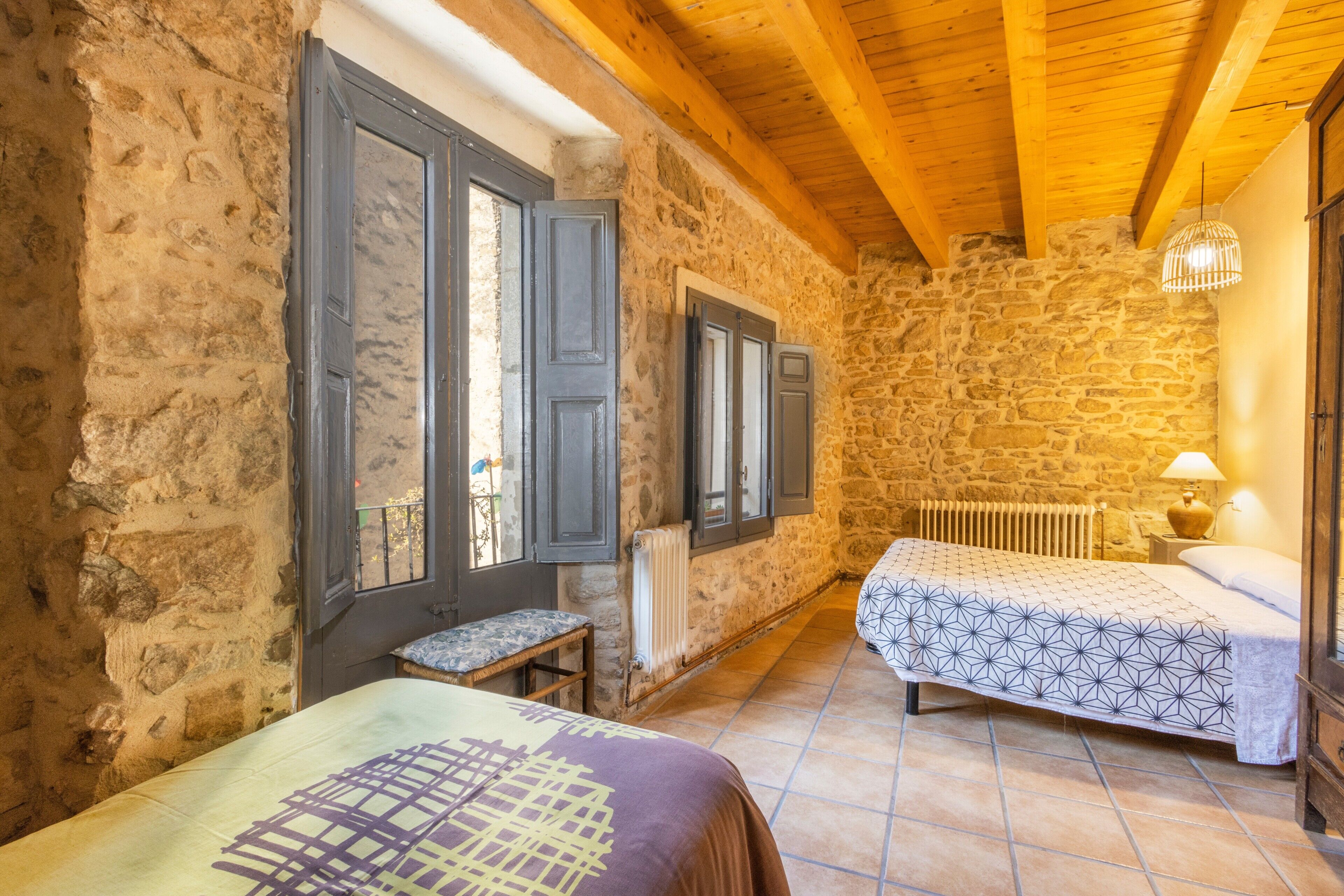 A rustic room with stone walls, wooden beams, a bed with patterned bedding, a bench, and a radiator.