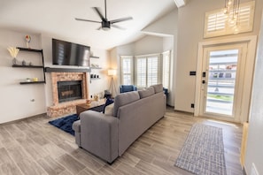 A modern living room with a fireplace, a grey sofa, a ceiling fan, and a large window with blinds. - Stylish Escape/3BDRM & fireplace (El Paso)