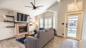 A modern living room with a fireplace, a grey sofa, a ceiling fan, and a large window with blinds. - Stylish Escape/3BDRM & fireplace (El Paso)