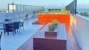 A rooftop terrace with wooden flooring, outdoor seating, and planter boxes with flowers. - Melhores Flats - Gracioso a 100m do mar (João Pessoa)