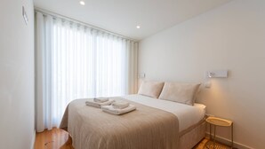 A neatly made bed with a beige bedspread and pillows, a small bedside table with a woven tray, and a window with sheer curtains allowing natural light. - Porto Luxe Stay: Garden & Pool (Porto)