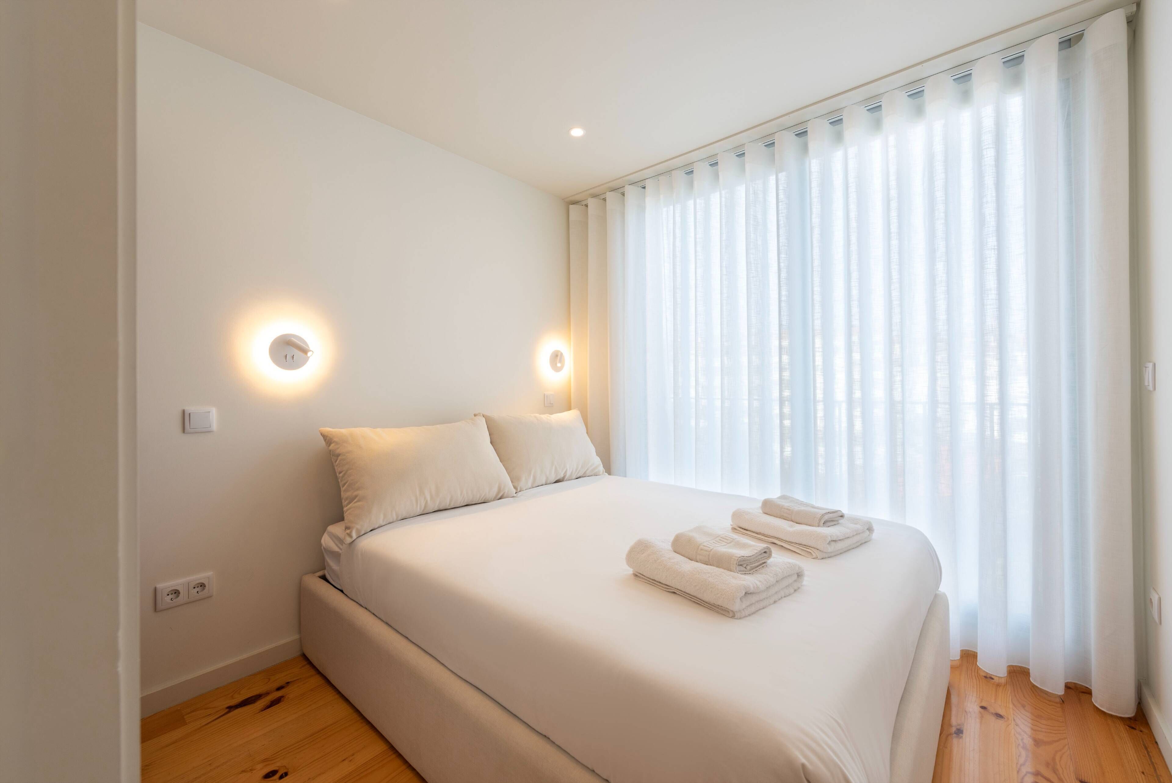 Una cama bien tendida con sábanas blancas y dos almohadas, una mesita de noche con lámpara y una ventana con cortinas traslúcidas.