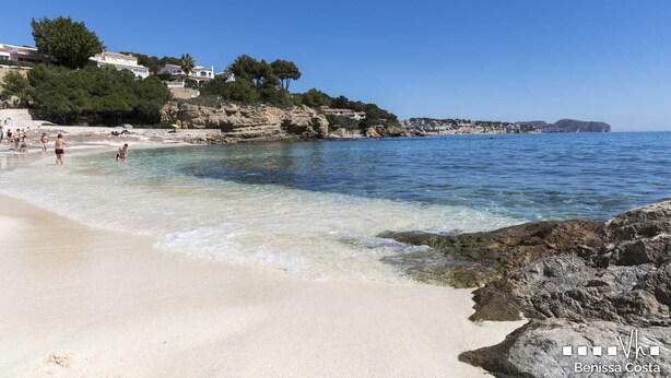 En strand med klart vatten, en stenig kust och byggnader som ligger på en sluttning.