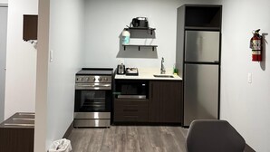 A compact kitchen with a stove, oven, microwave, and refrigerator. There is a dining table with chairs, a trash bin, and a fire extinguisher on the wall. - Zero-100 Motor Inn (Marathon)